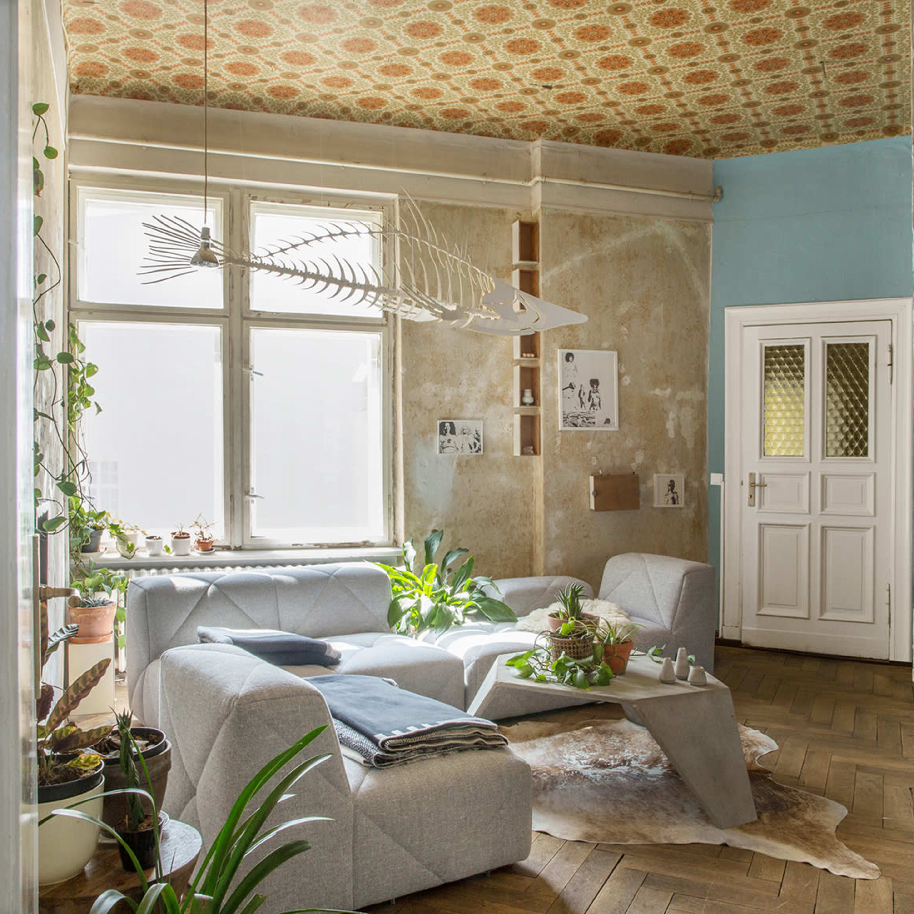 A light-filled living room features a gray sectional sofa, a minimalist coffee table, potted plants, and a decorative fish skeleton hanging from the ceiling.