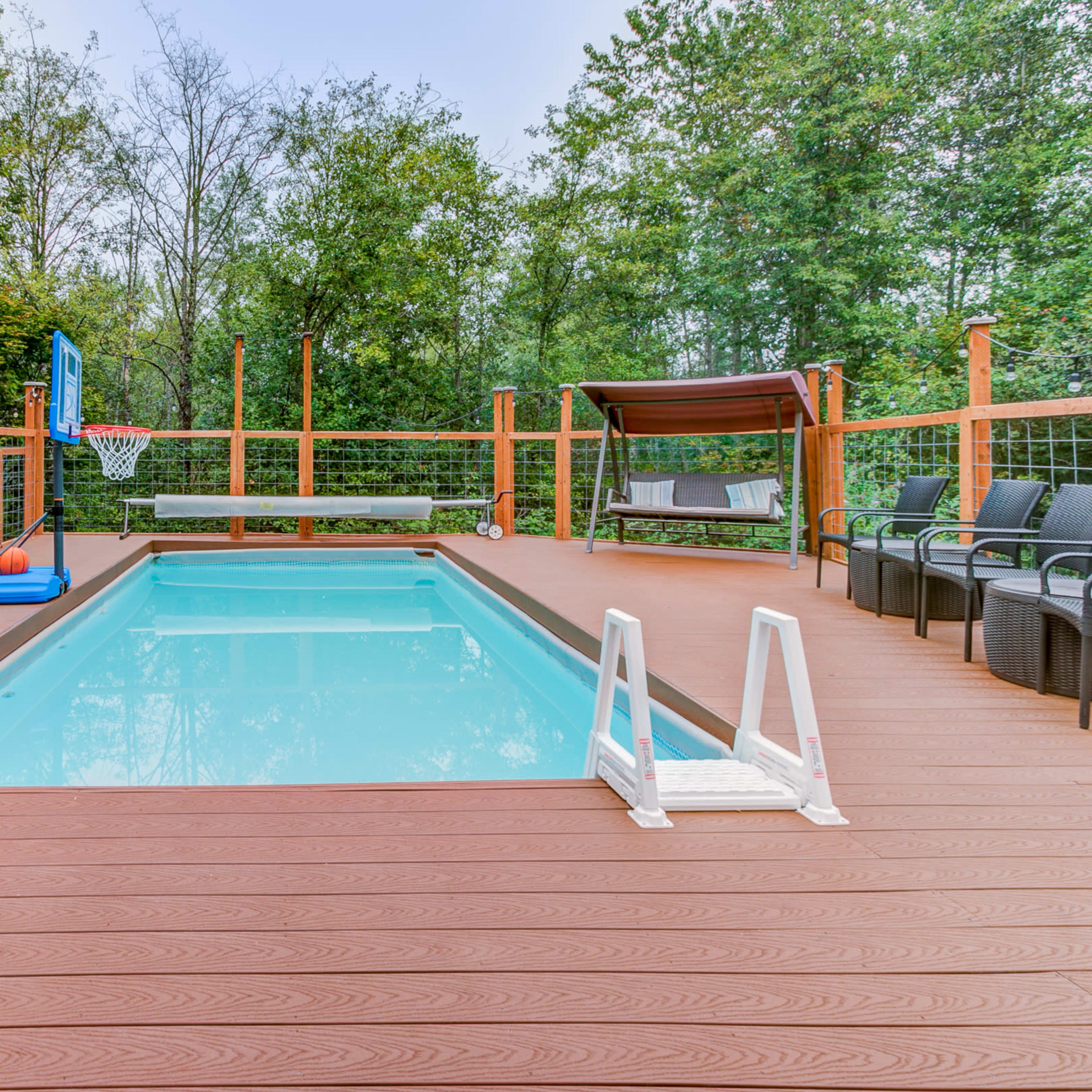 A swimming pool surrounded by a wooden deck, with a basketball hoop, seating area, and a shaded canopy nearby.