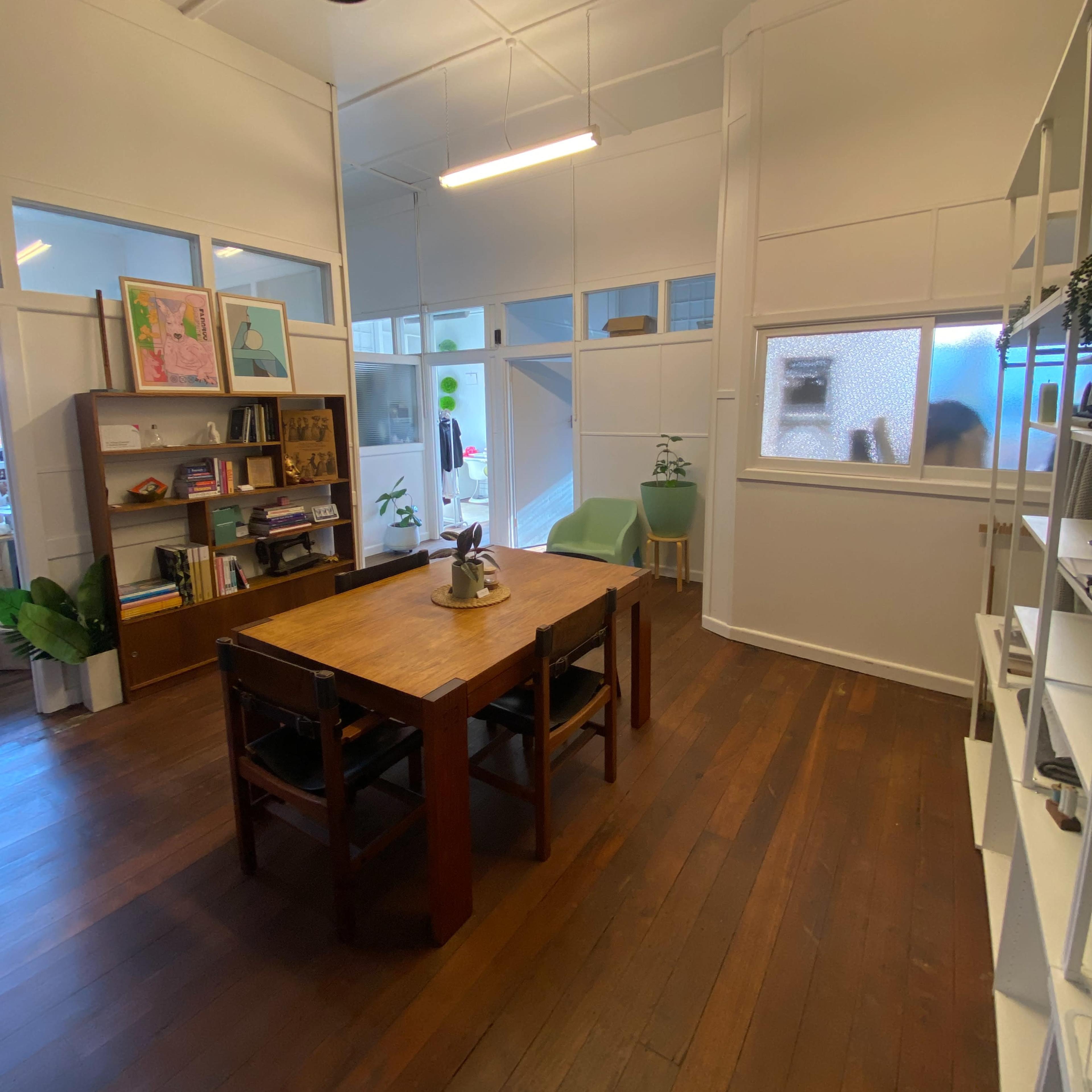 A bright room with wooden flooring, featuring a central table surrounded by chairs, a bookshelf filled with items, and large windows allowing natural light.