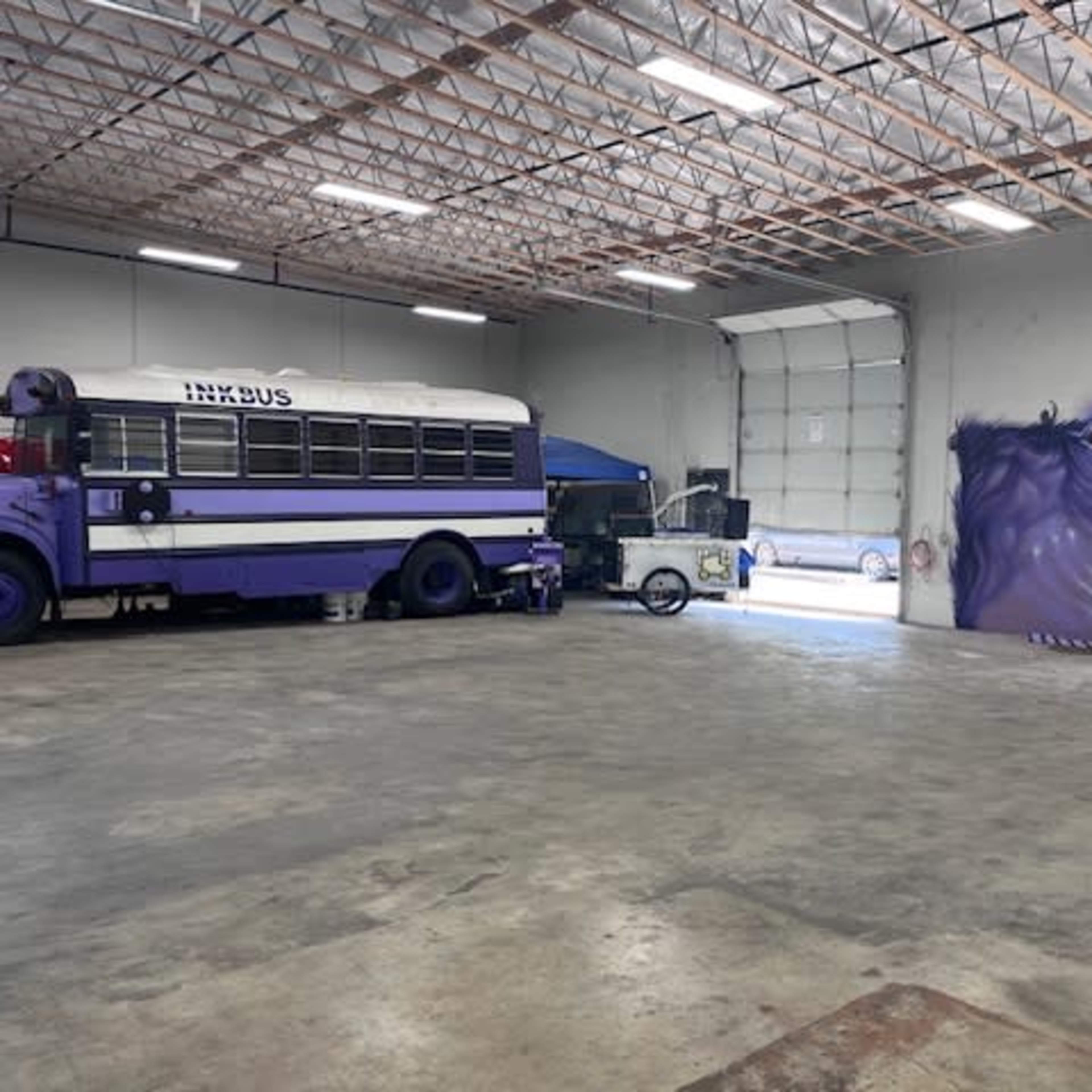 A purple school bus is parked inside a spacious industrial garage, with a mural of an eye on the gray wall.