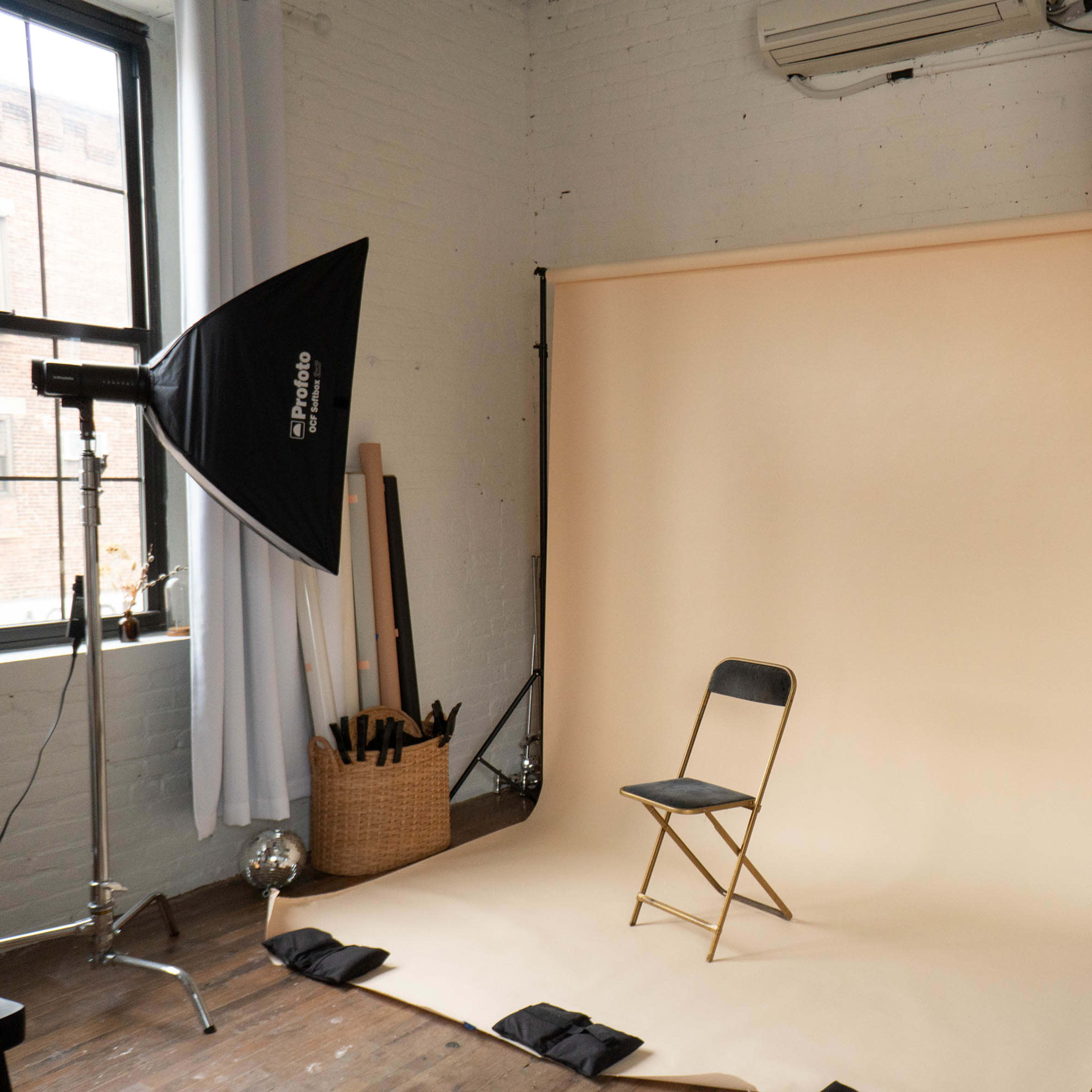 A photography setup features a empty folding chair in front of a neutral beige backdrop, with soft lighting equipment and decorative elements in a modern studio space.