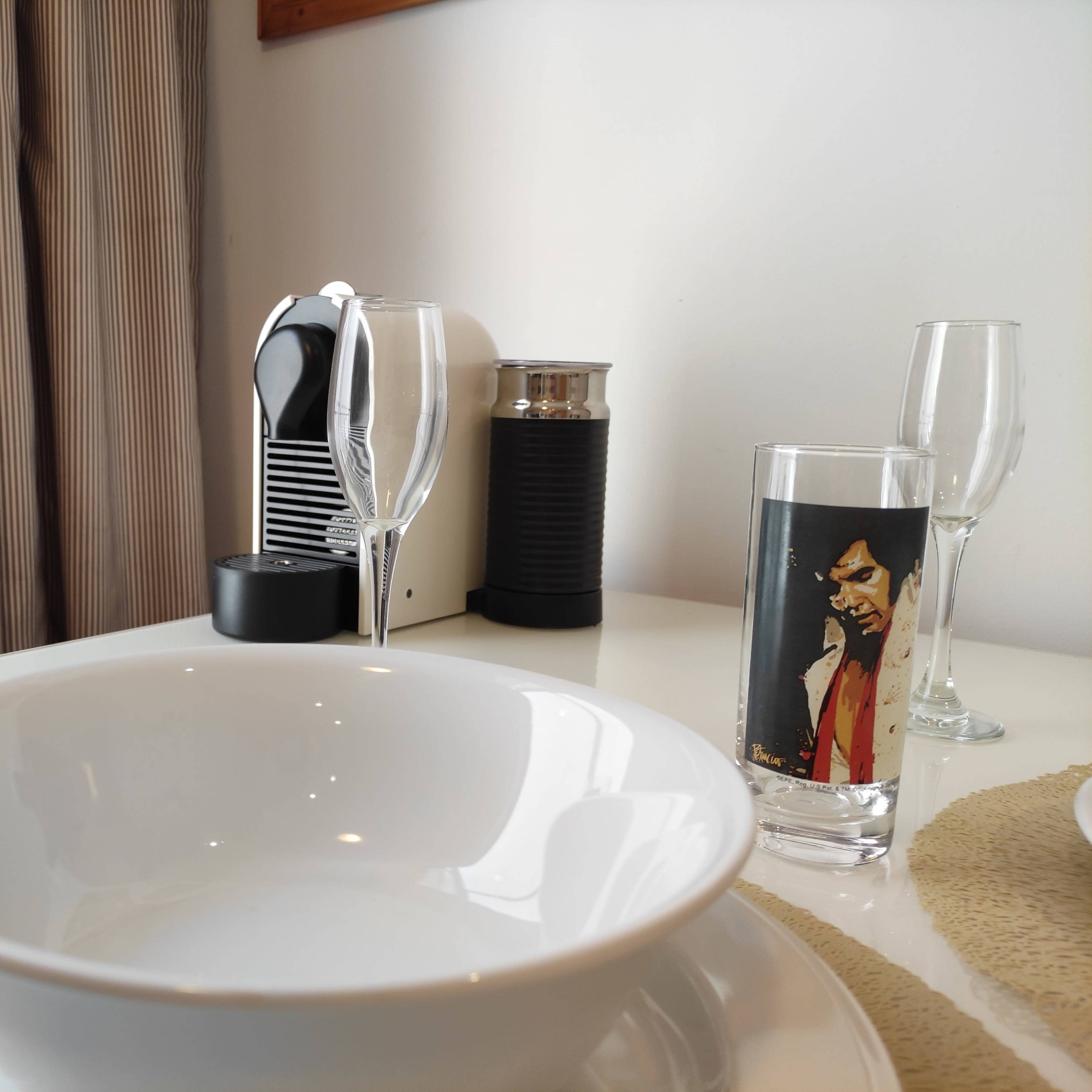 The scene shows a dining table set with a white bowl, two glasses featuring printed images, a coffee machine, and a pair of champagne flutes.