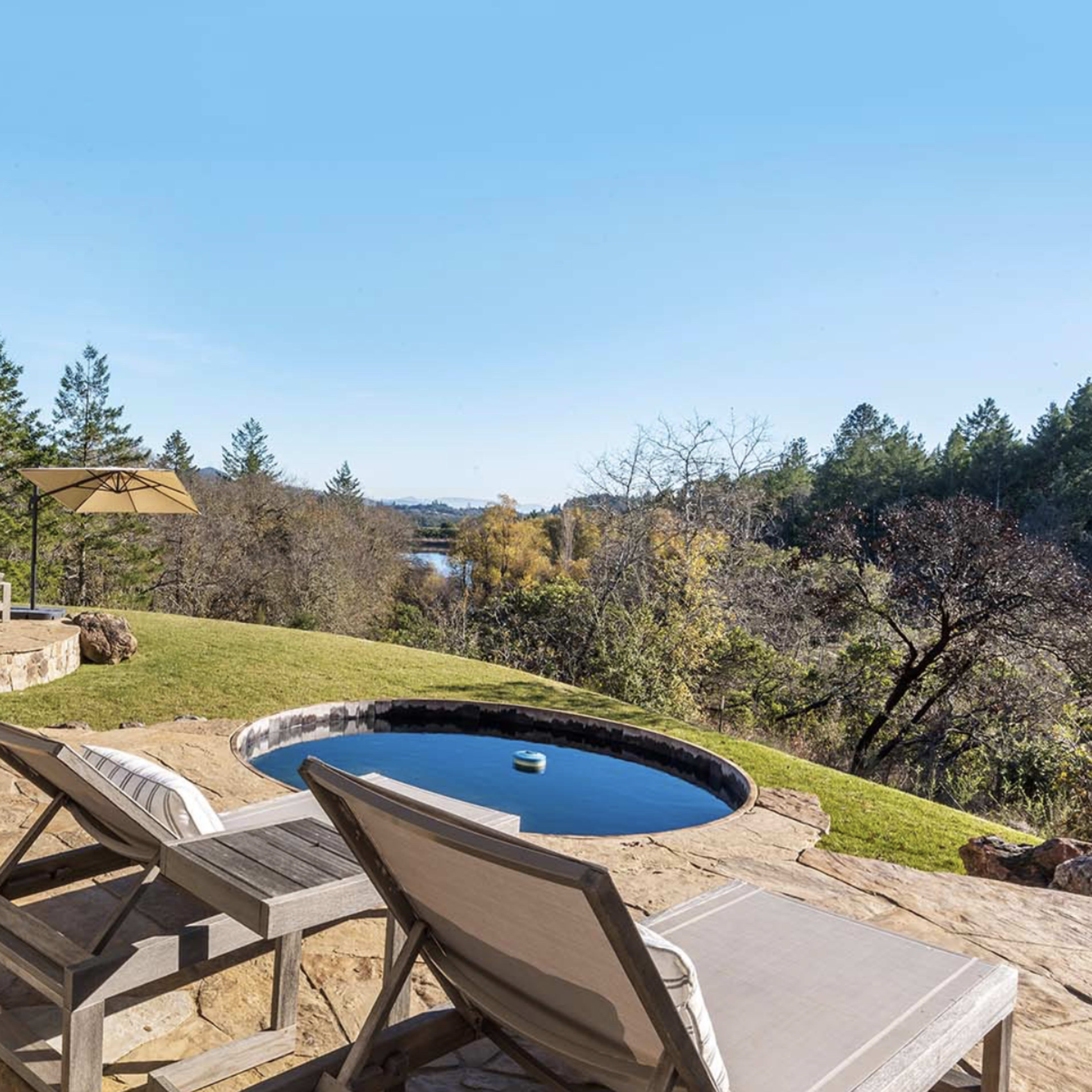 A backyard with a stone patio, a small circular hot tub, lounge chairs, and a view of a river and surrounding trees.