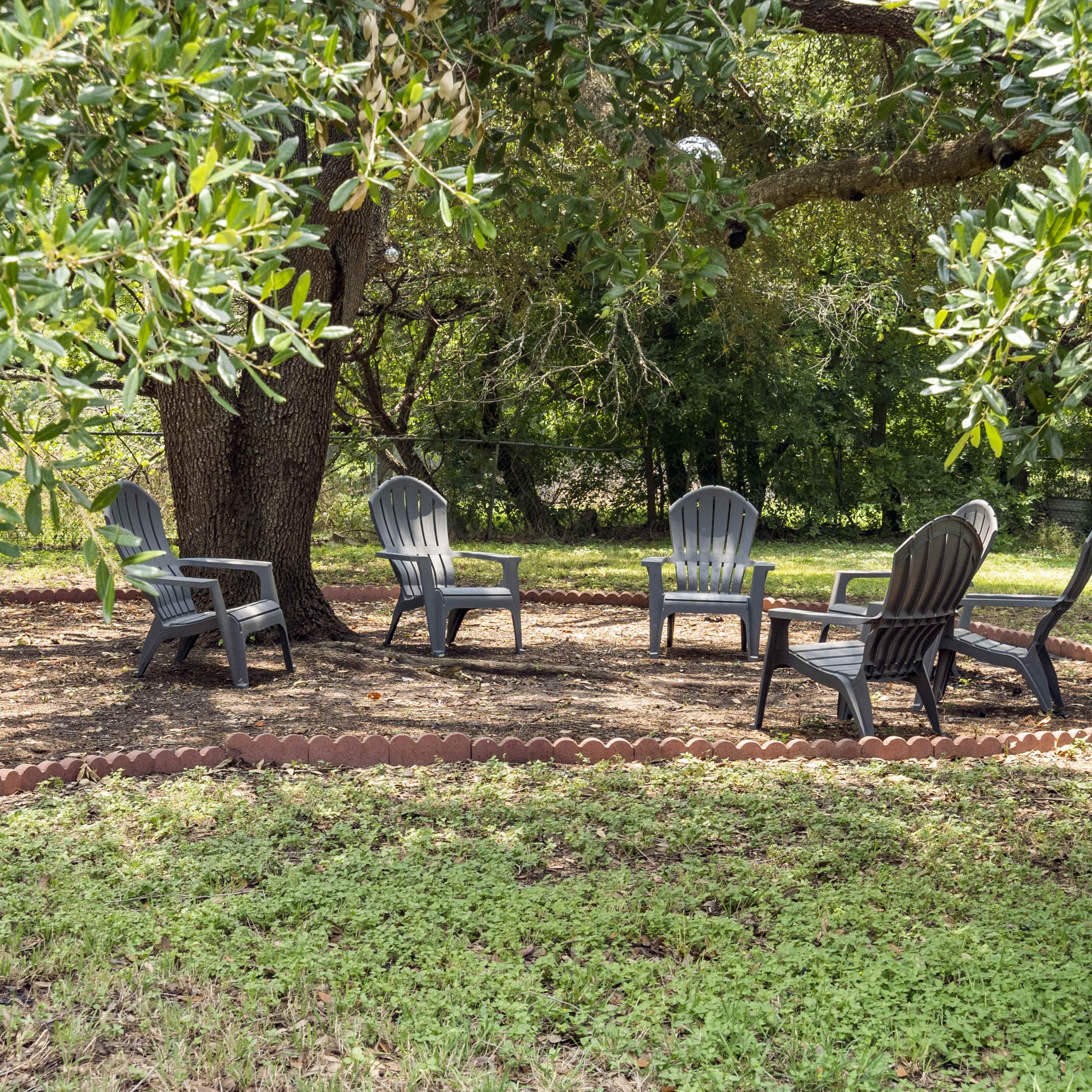 A circle of five gray adirondack chairs is arranged around a tree in a grassy area.