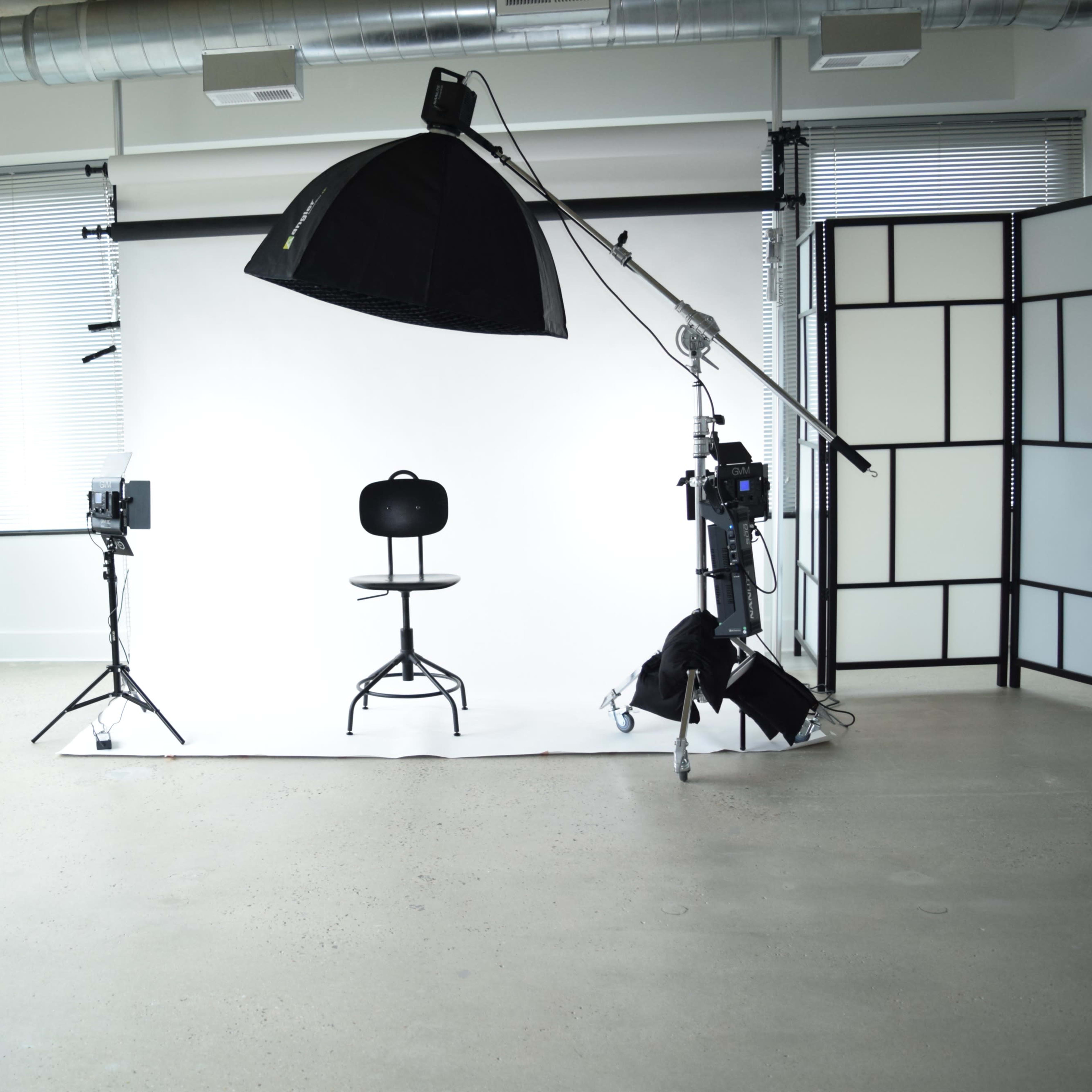 A photography studio featuring a white backdrop, a black stool, studio lighting equipment, and large windows with greenery outside.