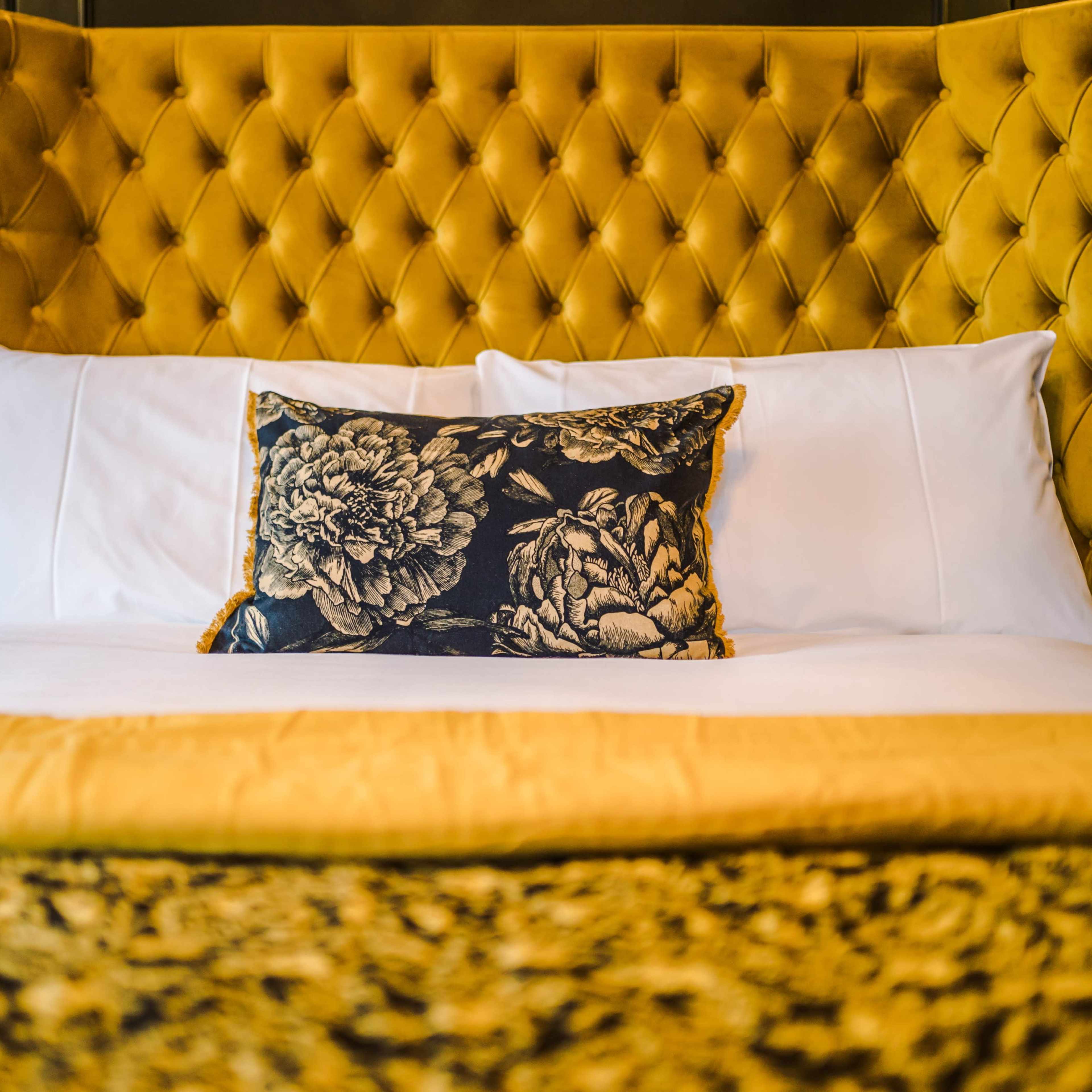 A neatly made bed with a tufted yellow headboard, white sheets, and a decorative black and gold floral pillow.