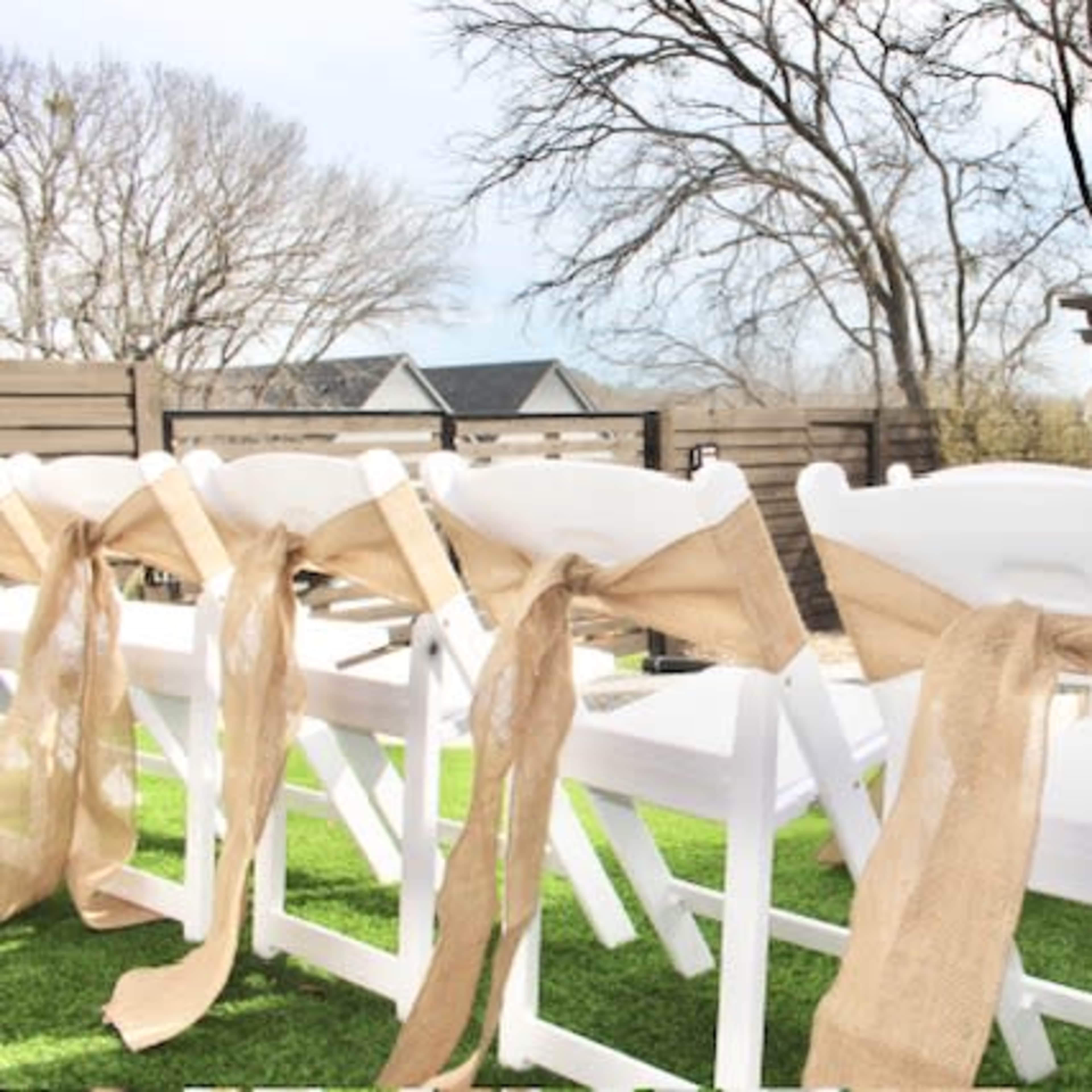 A set of white chairs arranged in an outdoor setting, adorned with burlap bows, with trees and a gazebo visible in the background.