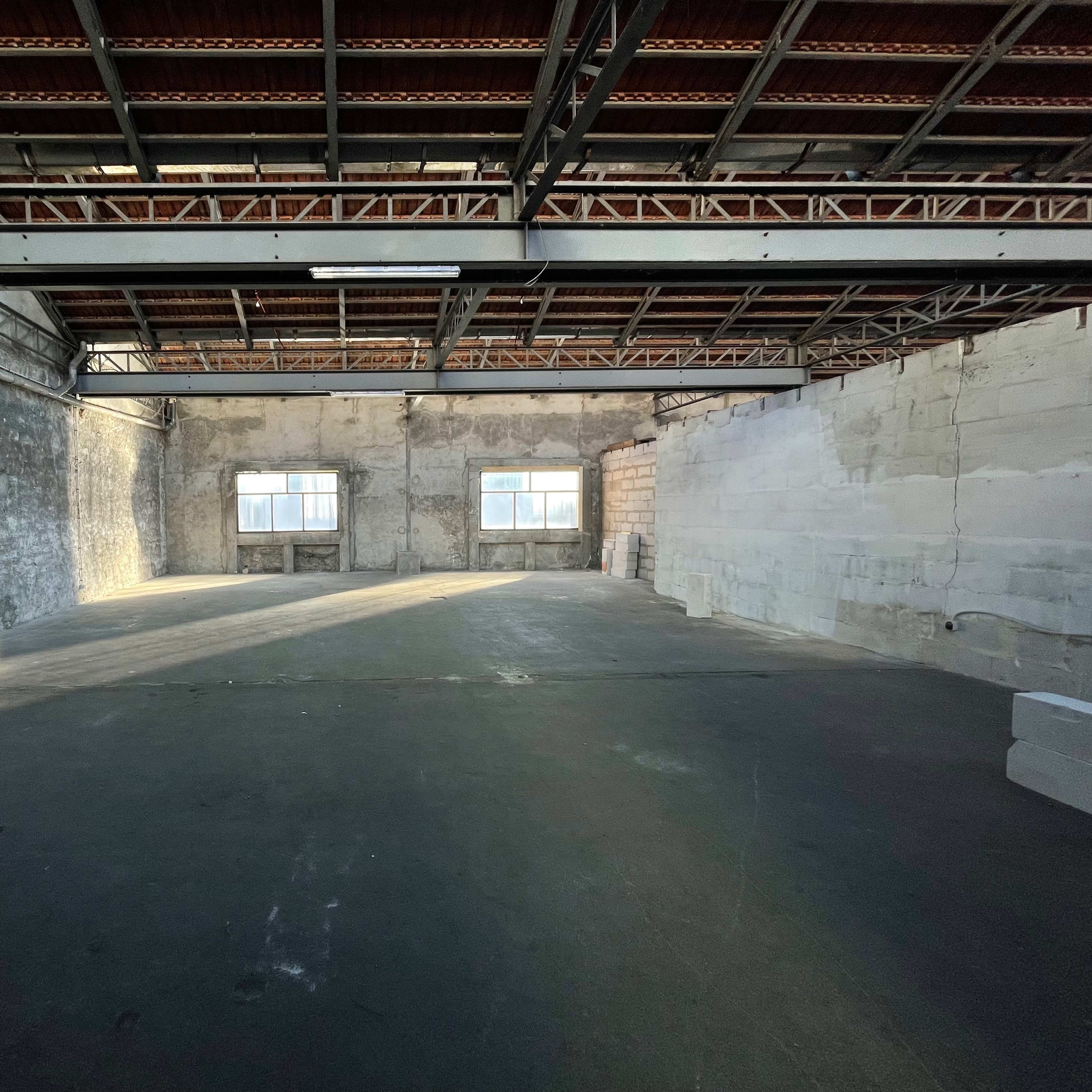 An empty industrial space with exposed brick walls, high ceilings, and two windows allowing natural light to enter.