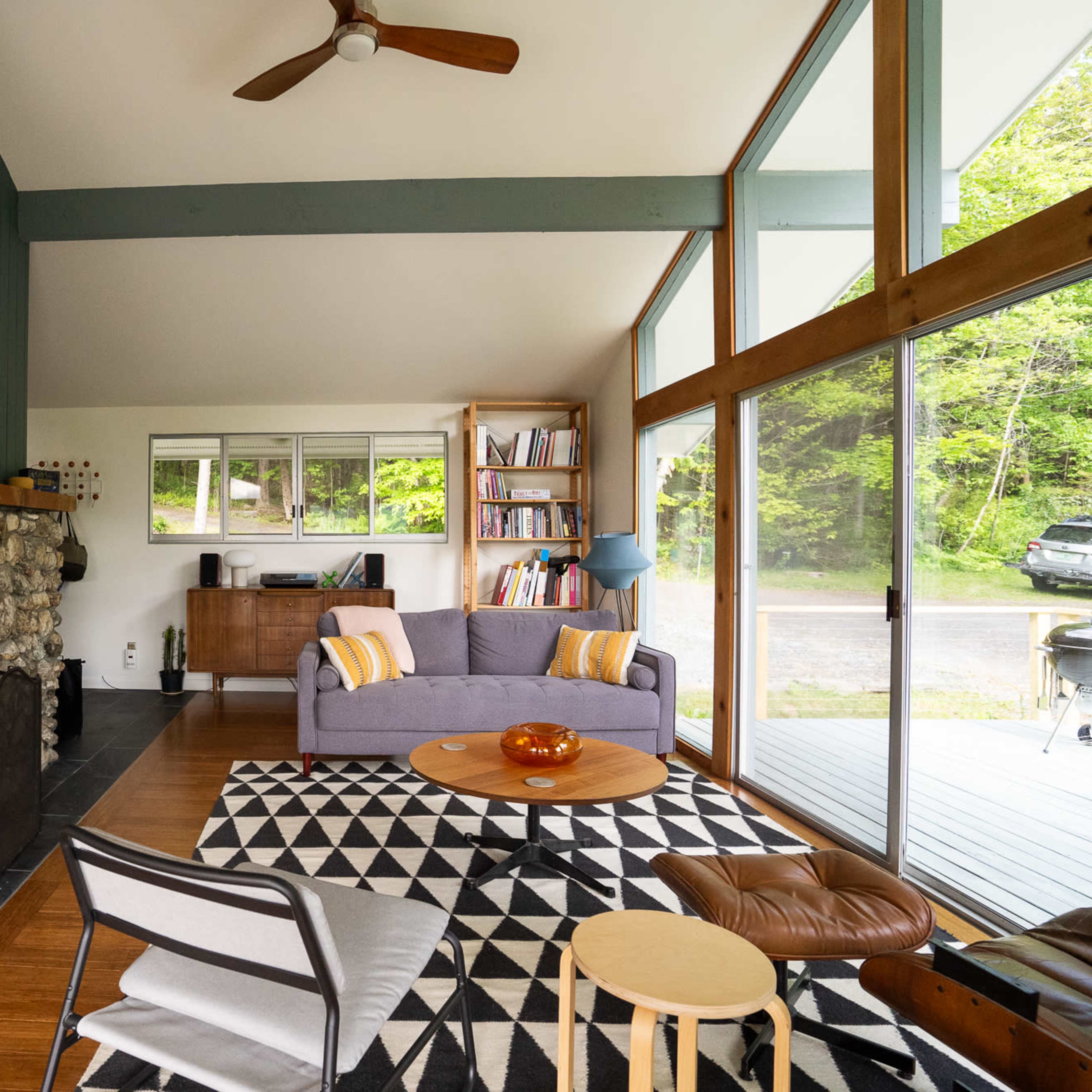 The interior of a modern living room features a stone fireplace, large windows, and a mix of furniture including a gray sofa and a round coffee table on a patterned rug.