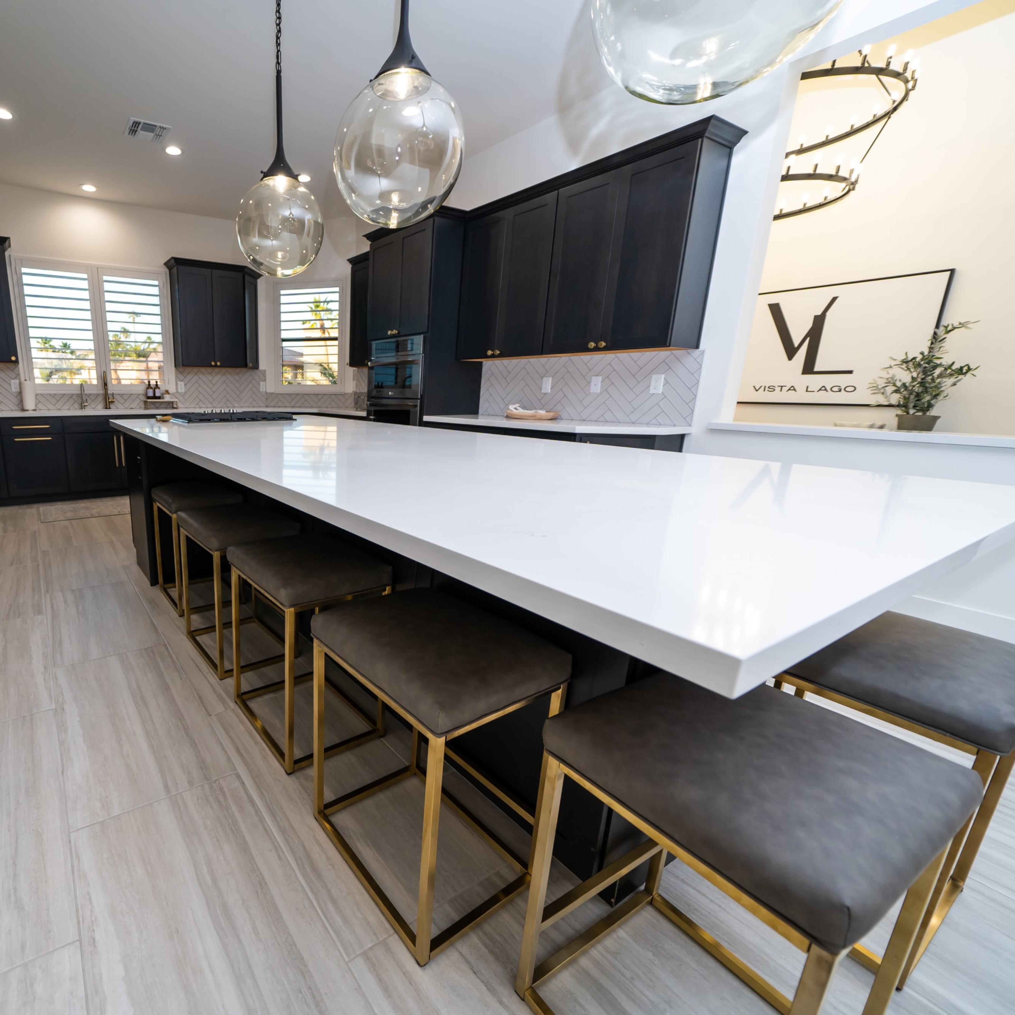 A modern kitchen features a large white countertop with gold-accented stools and dark cabinetry.