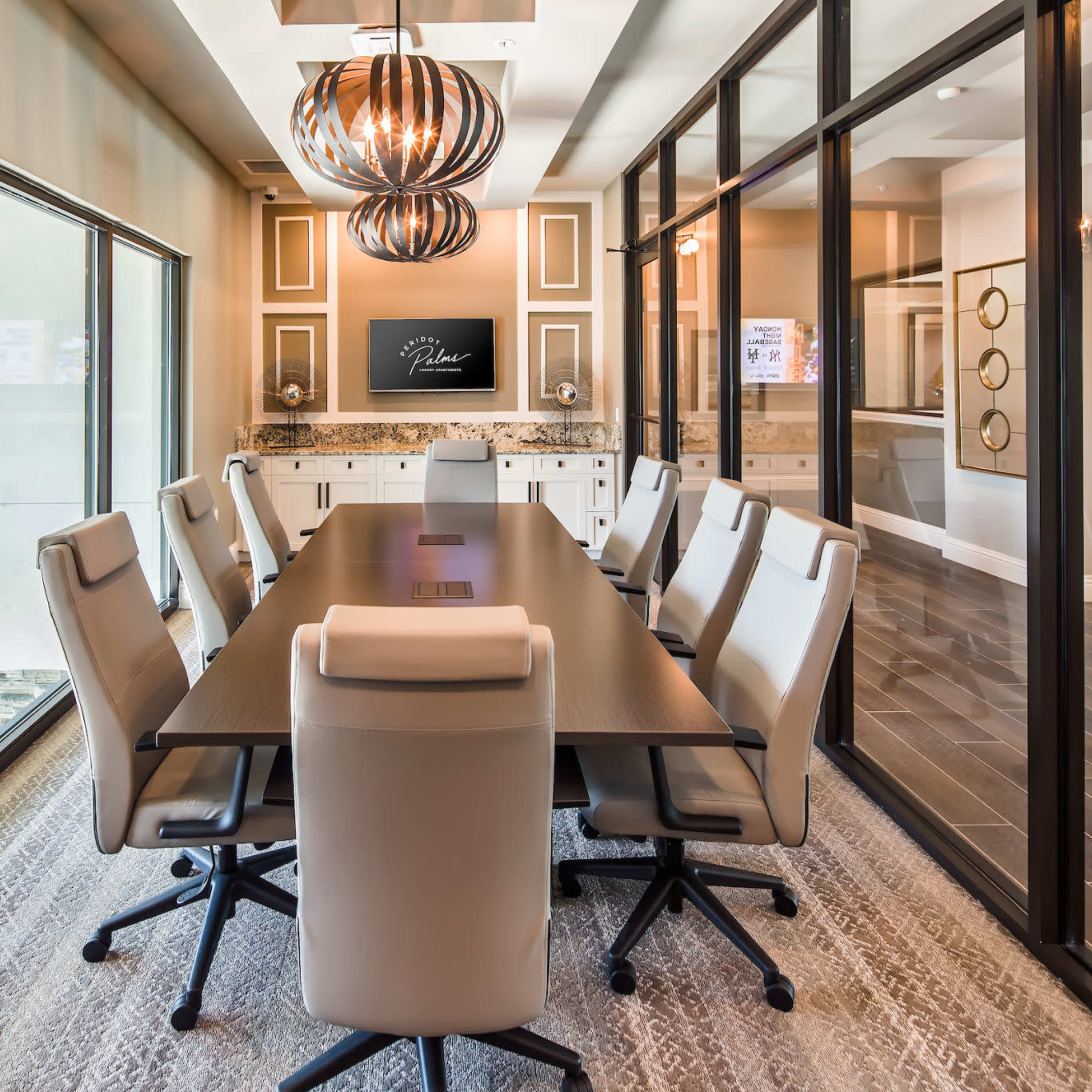 A modern conference room features a large table surrounded by white chairs, with glass walls and a decorative ceiling light.