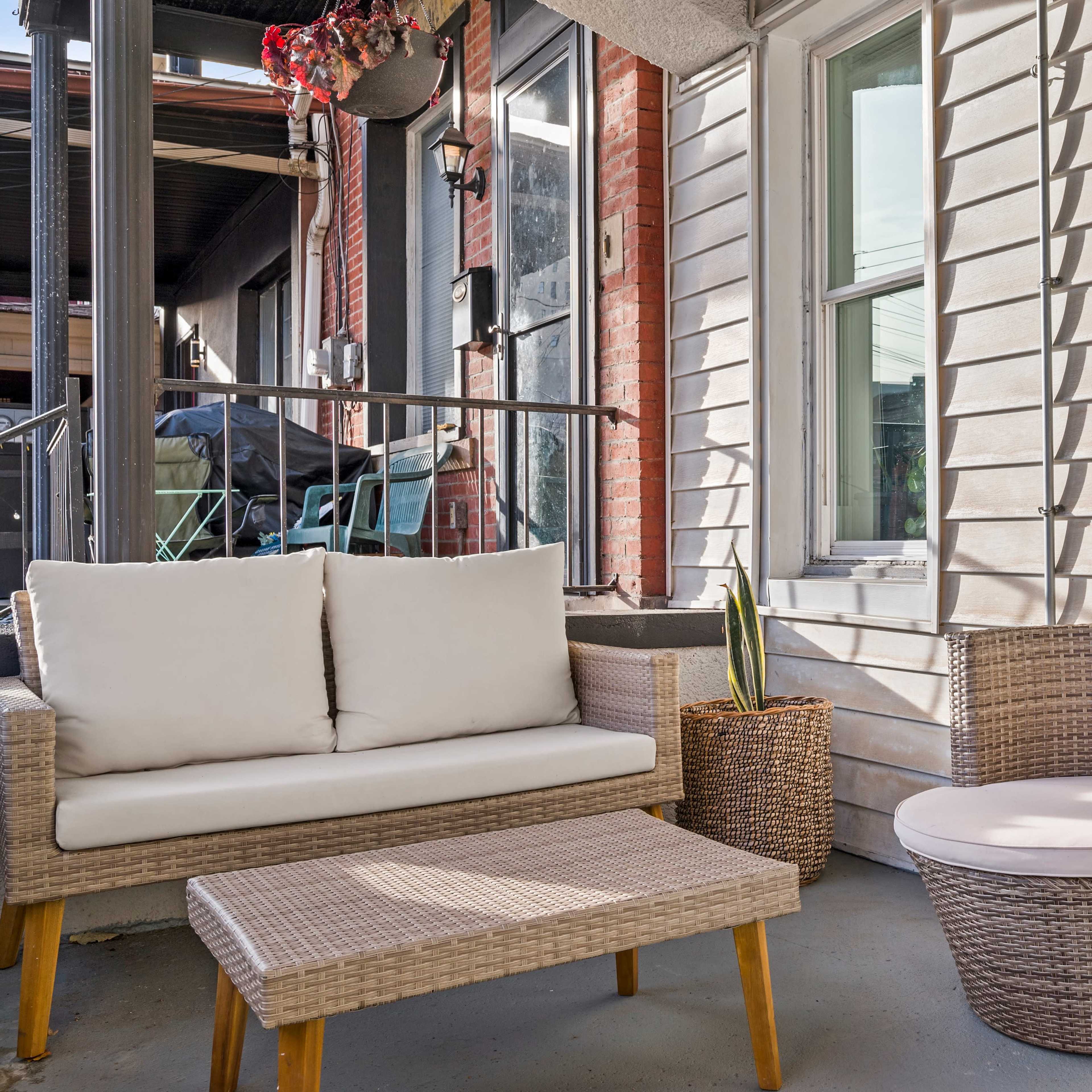 A small outdoor seating area featuring a wicker sofa, a side table, and a round chair, surrounded by windows and brick walls.