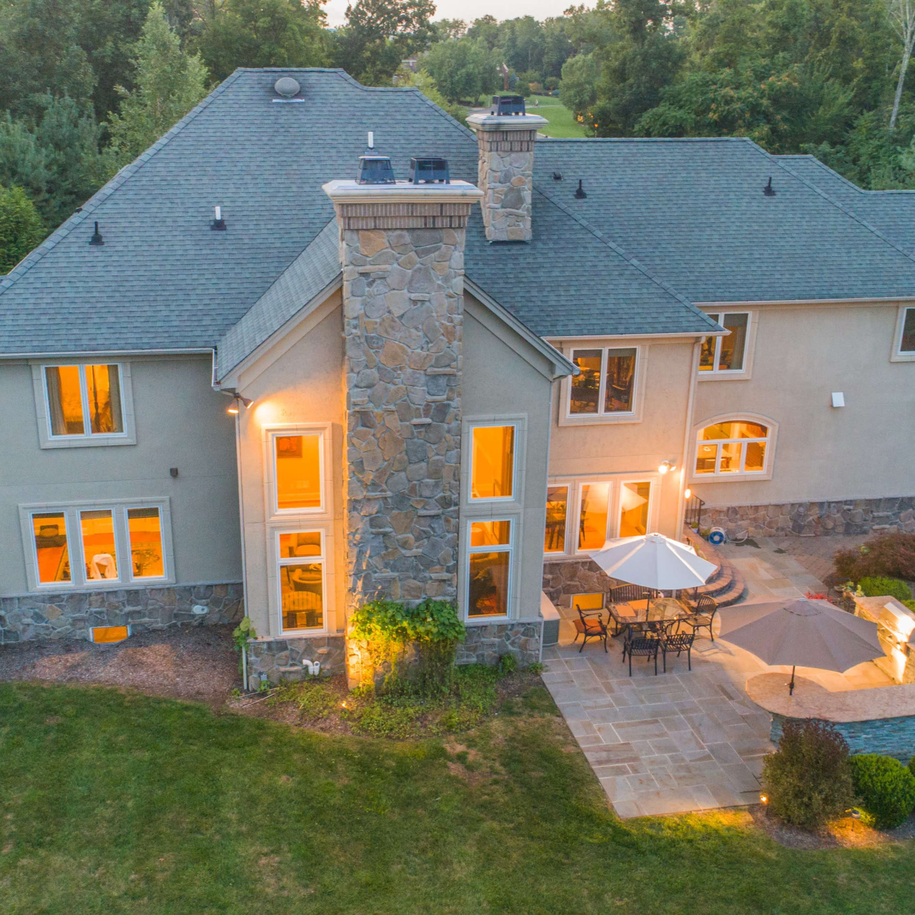 A large, contemporary home with a stone chimney and multiple windows is surrounded by a manicured lawn and a stone patio.