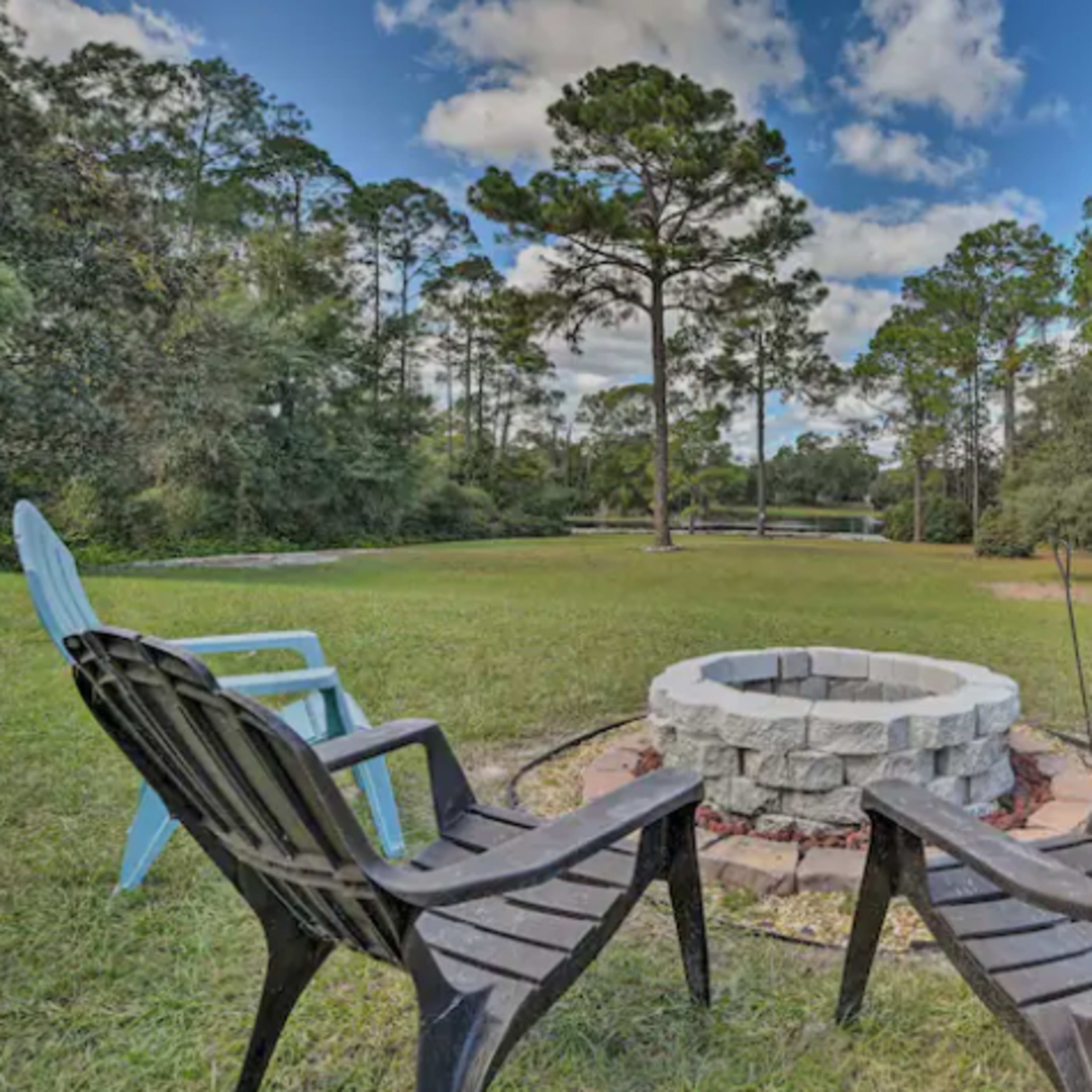 A circular stone fire pit is surrounded by four chairs on a grassy area near a body of water and tall trees.