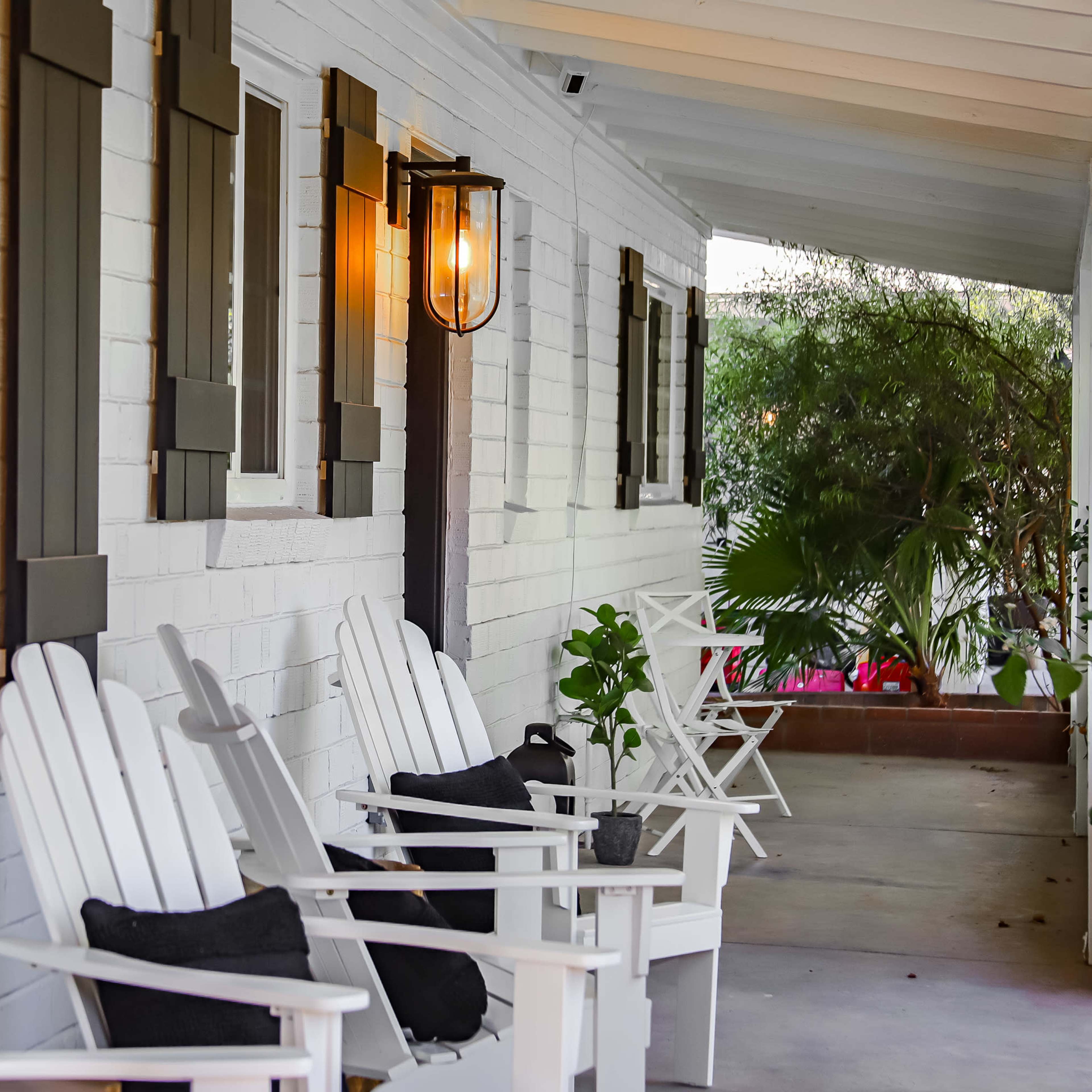 A porch with white wooden chairs arranged beside a wall adorned with windows and a lantern, surrounded by greenery.