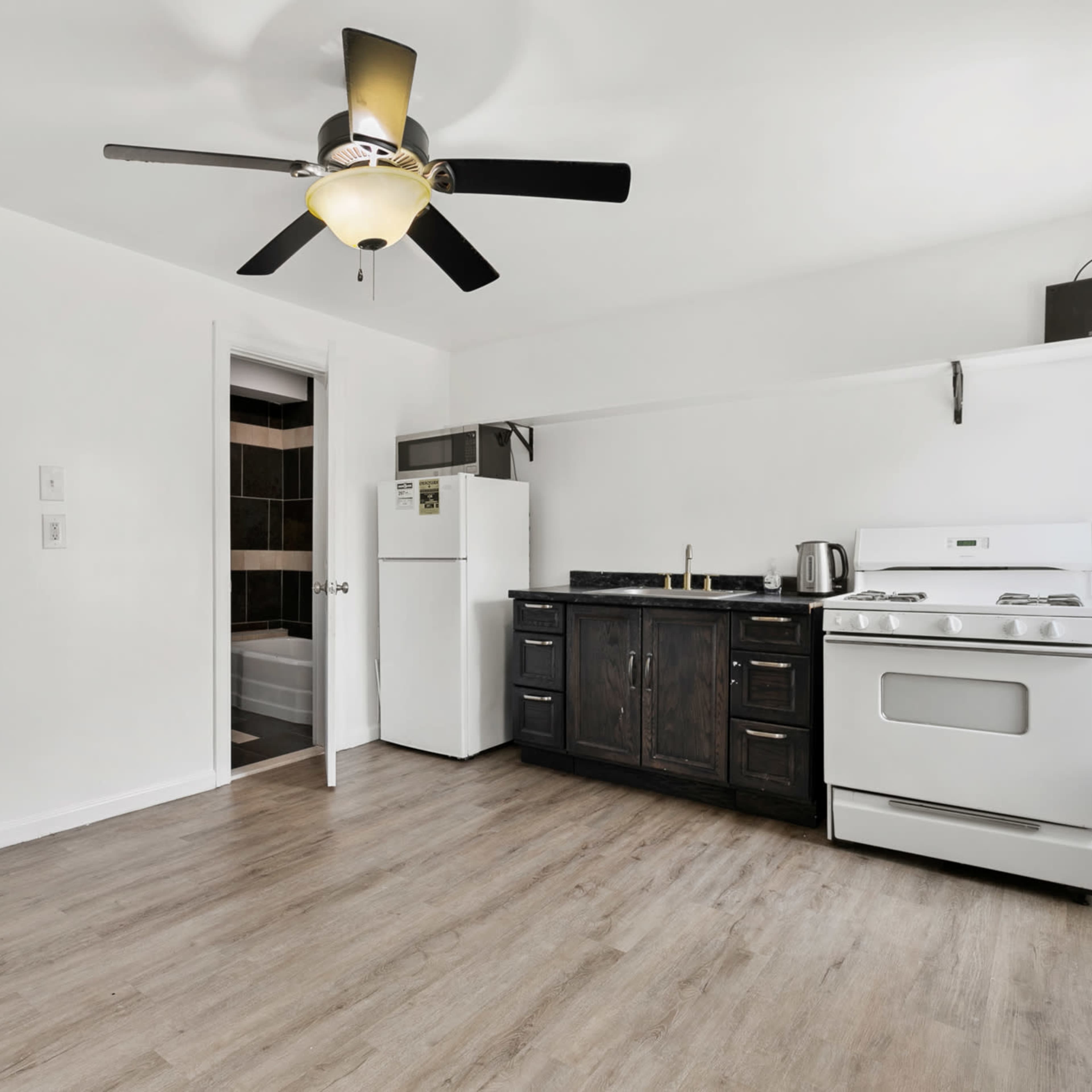 A simple kitchen with a white ceiling fan, a white refrigerator, a black kitchen cabinet, and a stove, all set against light wood flooring and white walls.