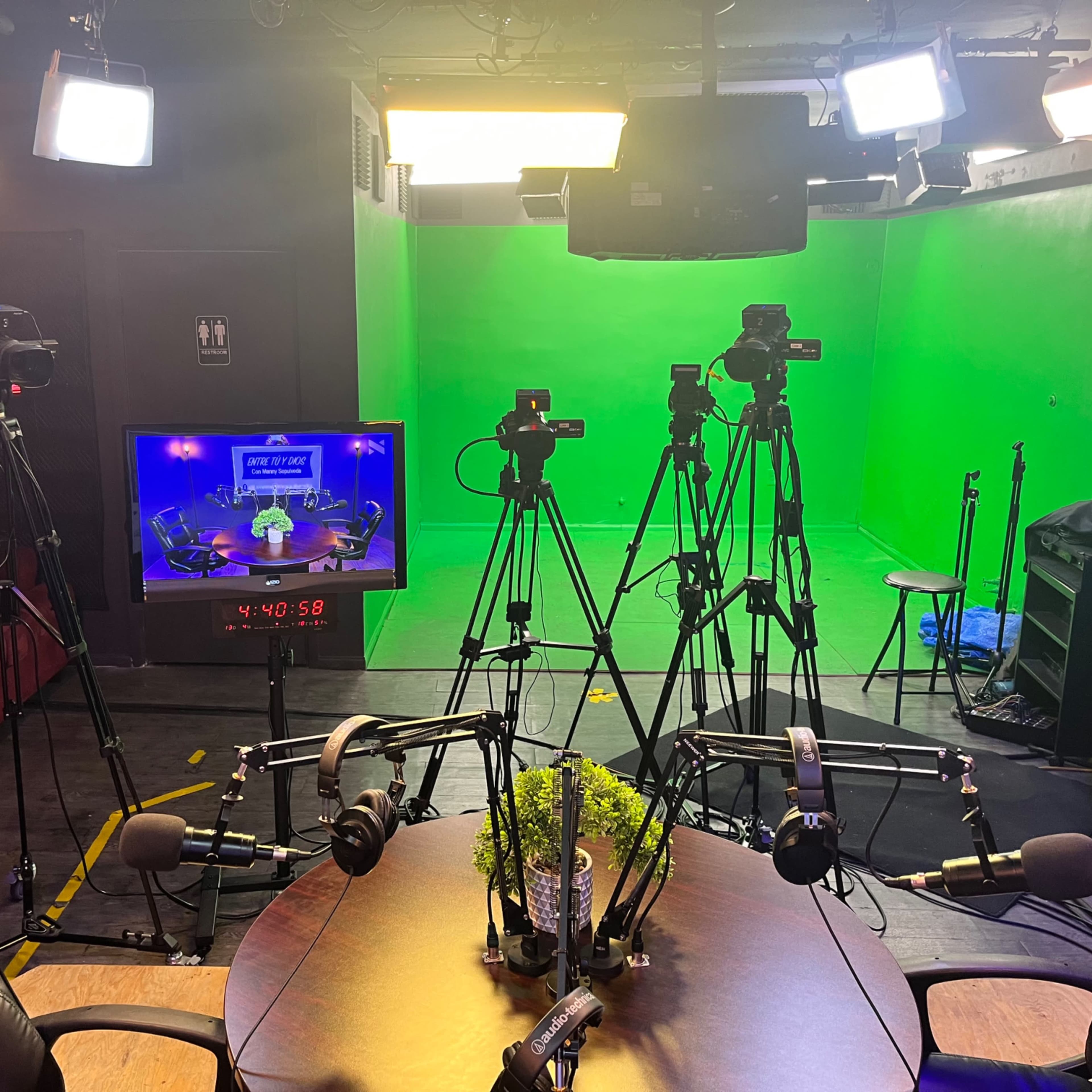 A television studio with multiple cameras, a green screen backdrop, and a central table equipped with several microphones.