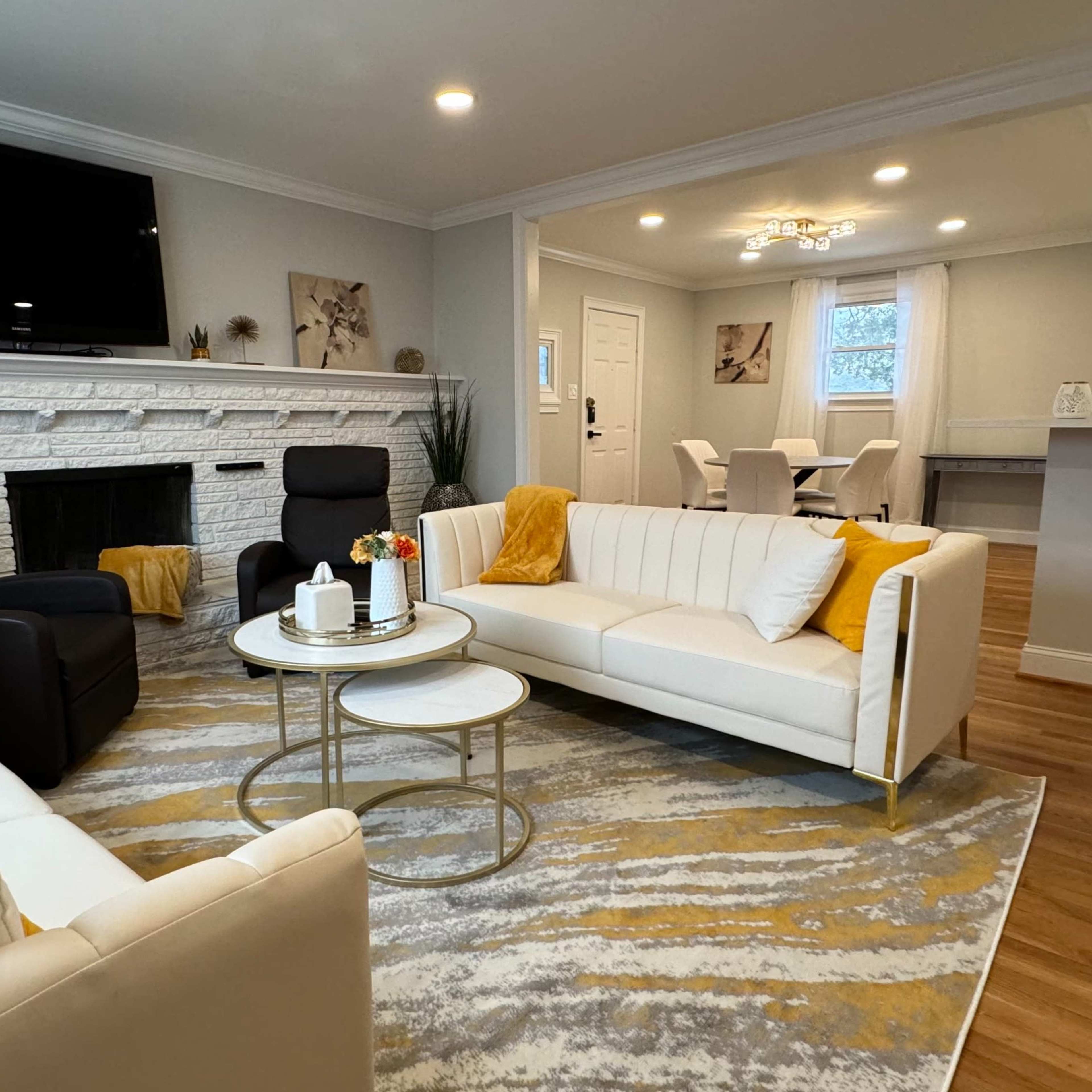 A living room with a white sofa, two armchairs, a circular coffee table, and a fireplace, all arranged around a patterned rug under overhead lighting.