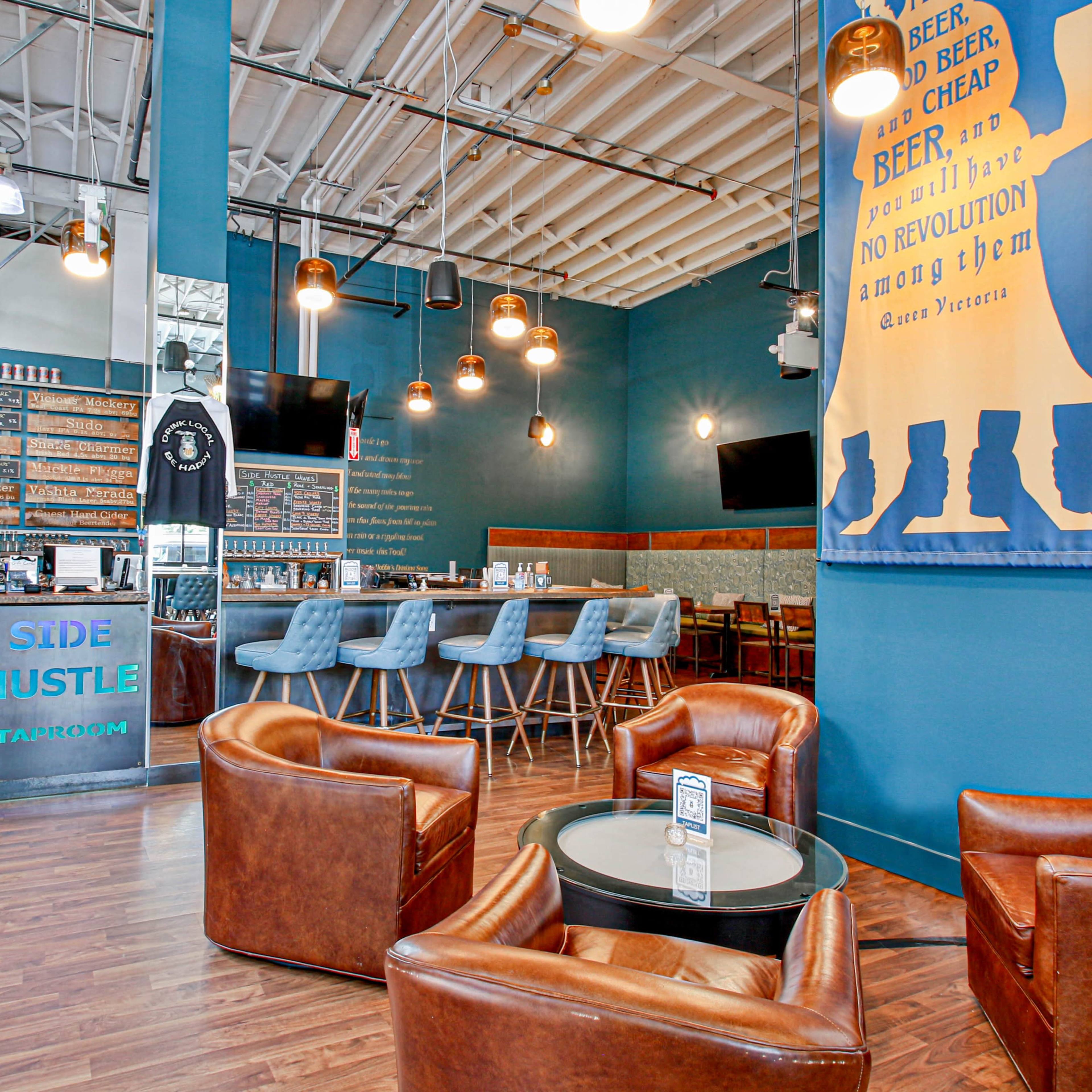 A modern taproom interior featuring a bar with high stools, wooden flooring, and leather chairs arranged around a table.