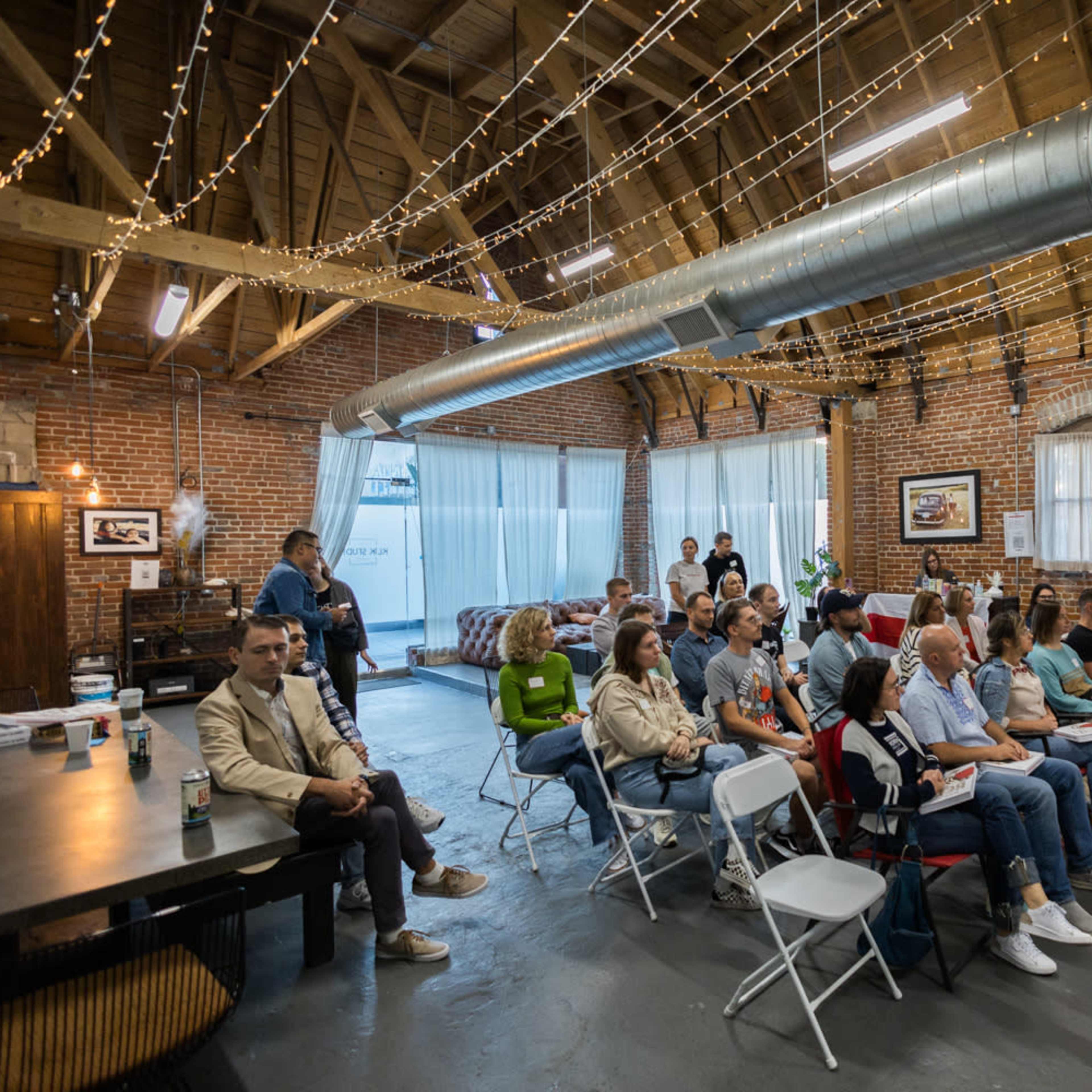 A group of people is seated in folding chairs within a spacious brick-walled venue, with decorative string lights overhead.