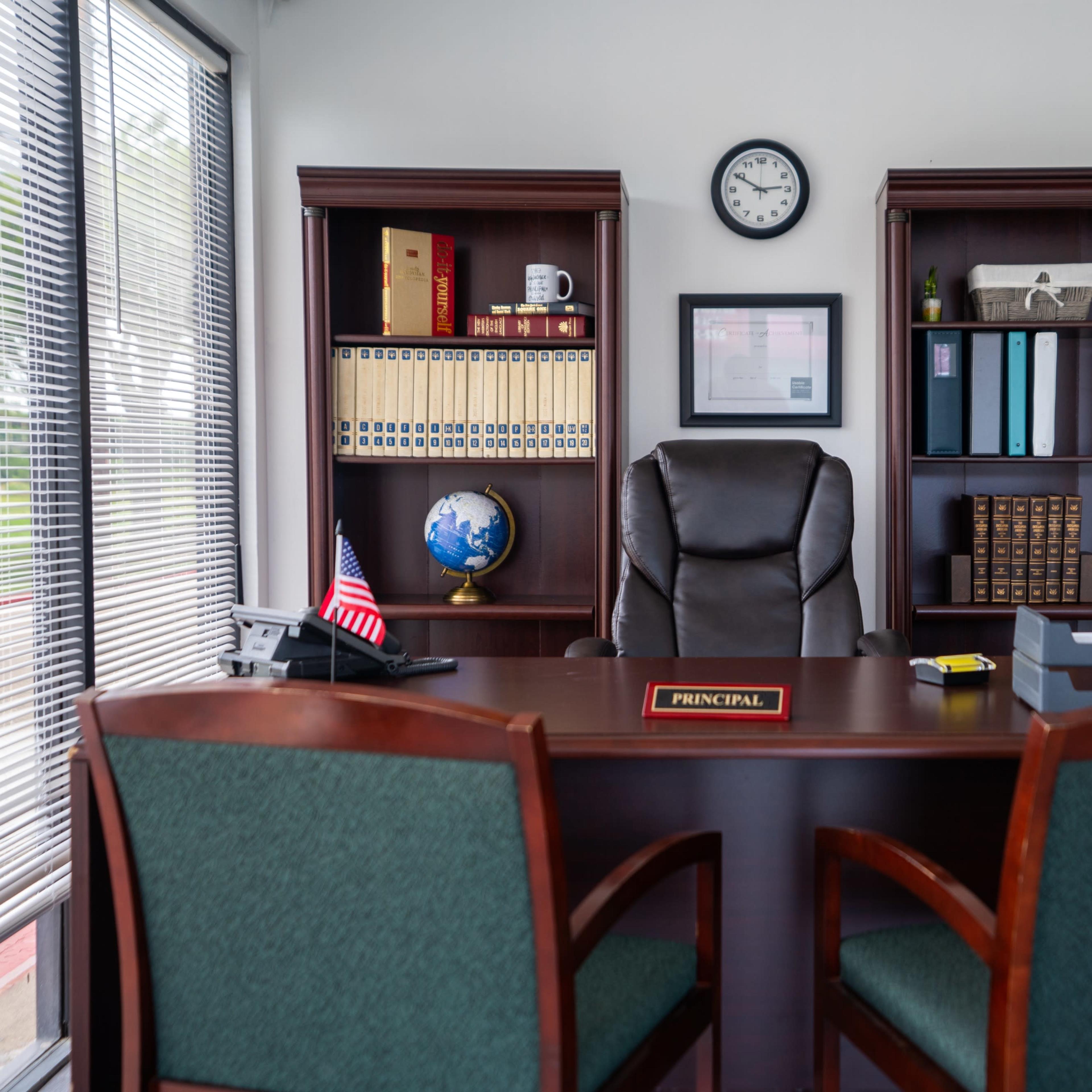 A classroom is arranged with empty wooden desks facing a green chalkboard and a teacher's desk at the front.