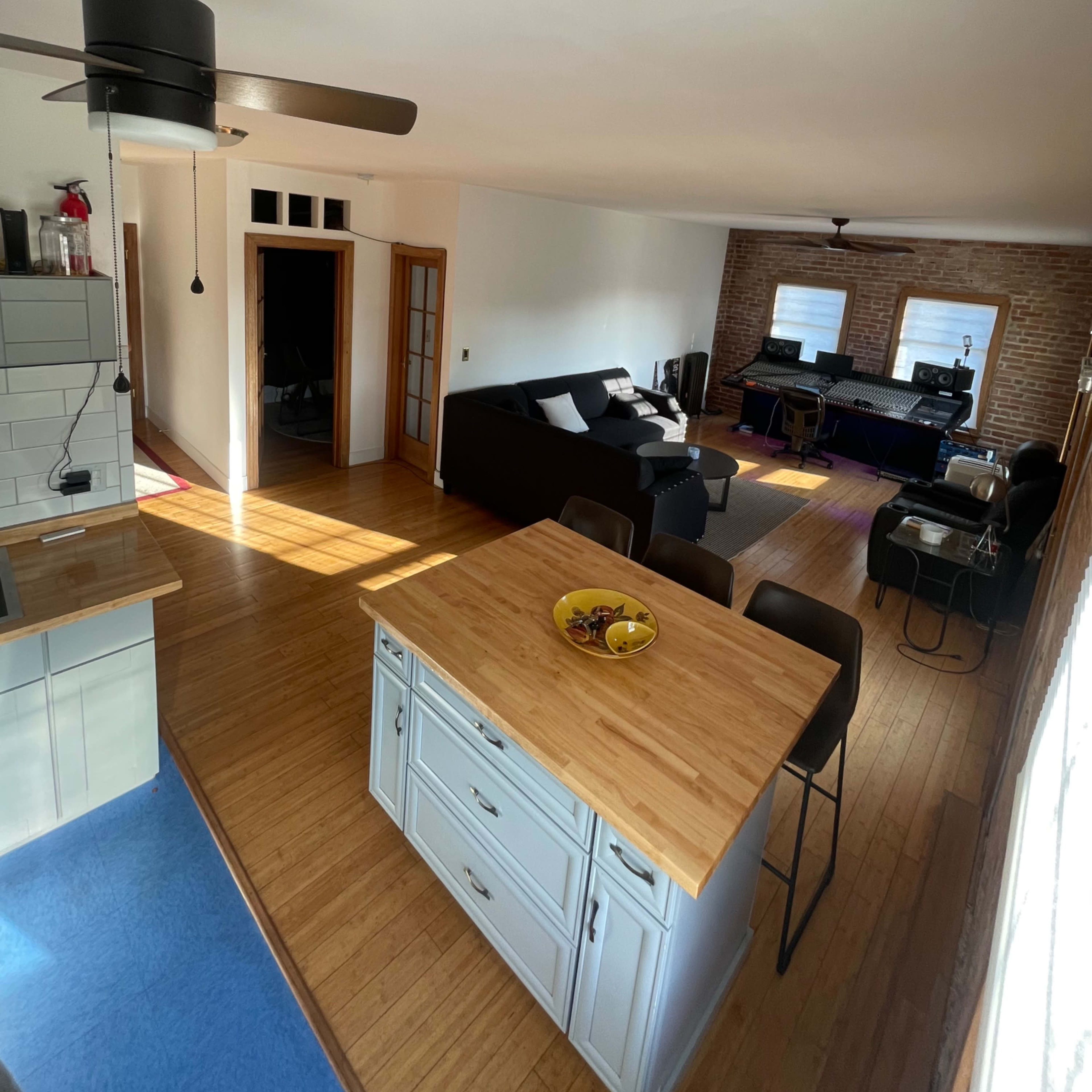 A bright living space features a kitchen island with a fruit bowl, a couch, and a workspace, all with wooden flooring and brick walls.