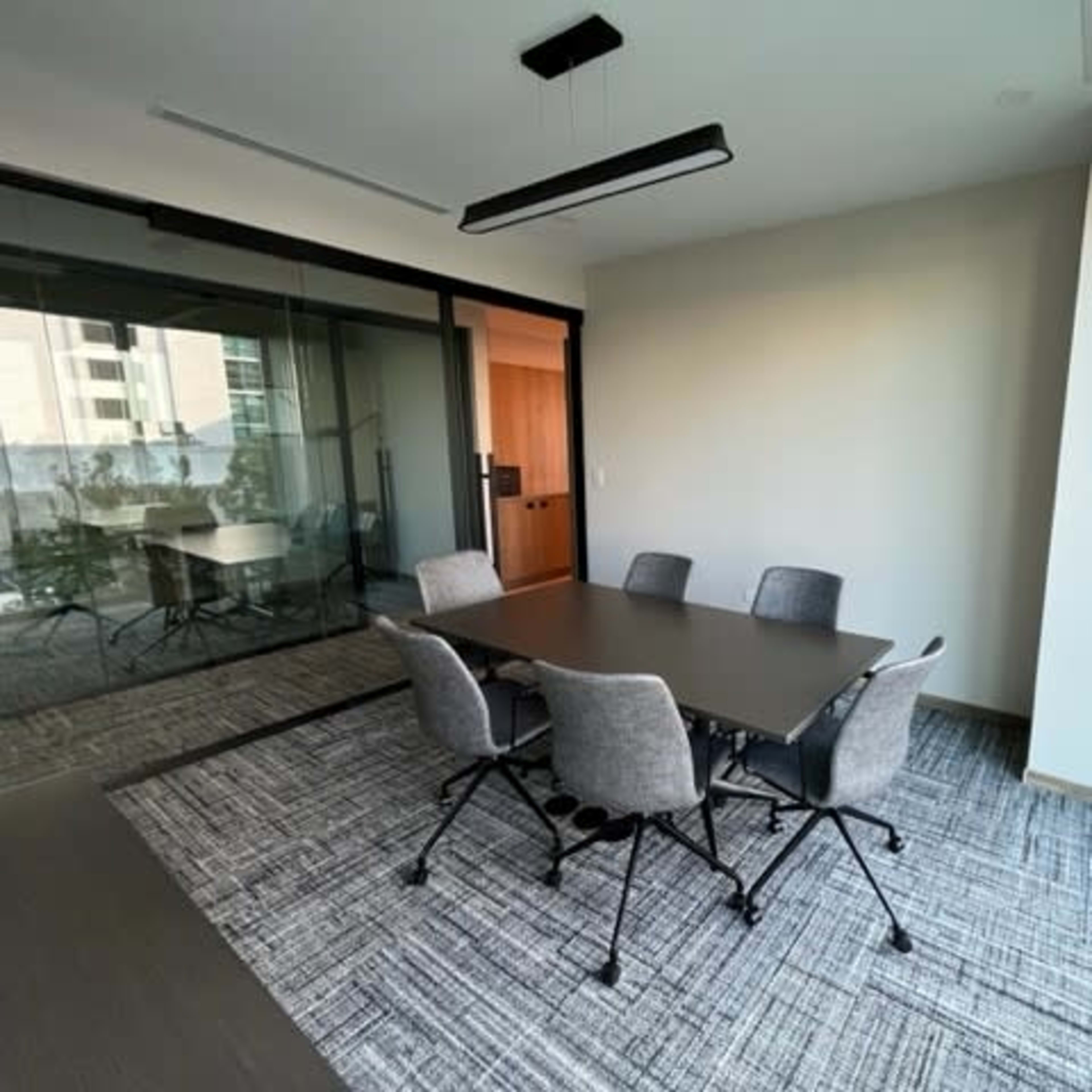 A conference room features a rectangular table surrounded by six chairs, with large windows offering a view of the outside.