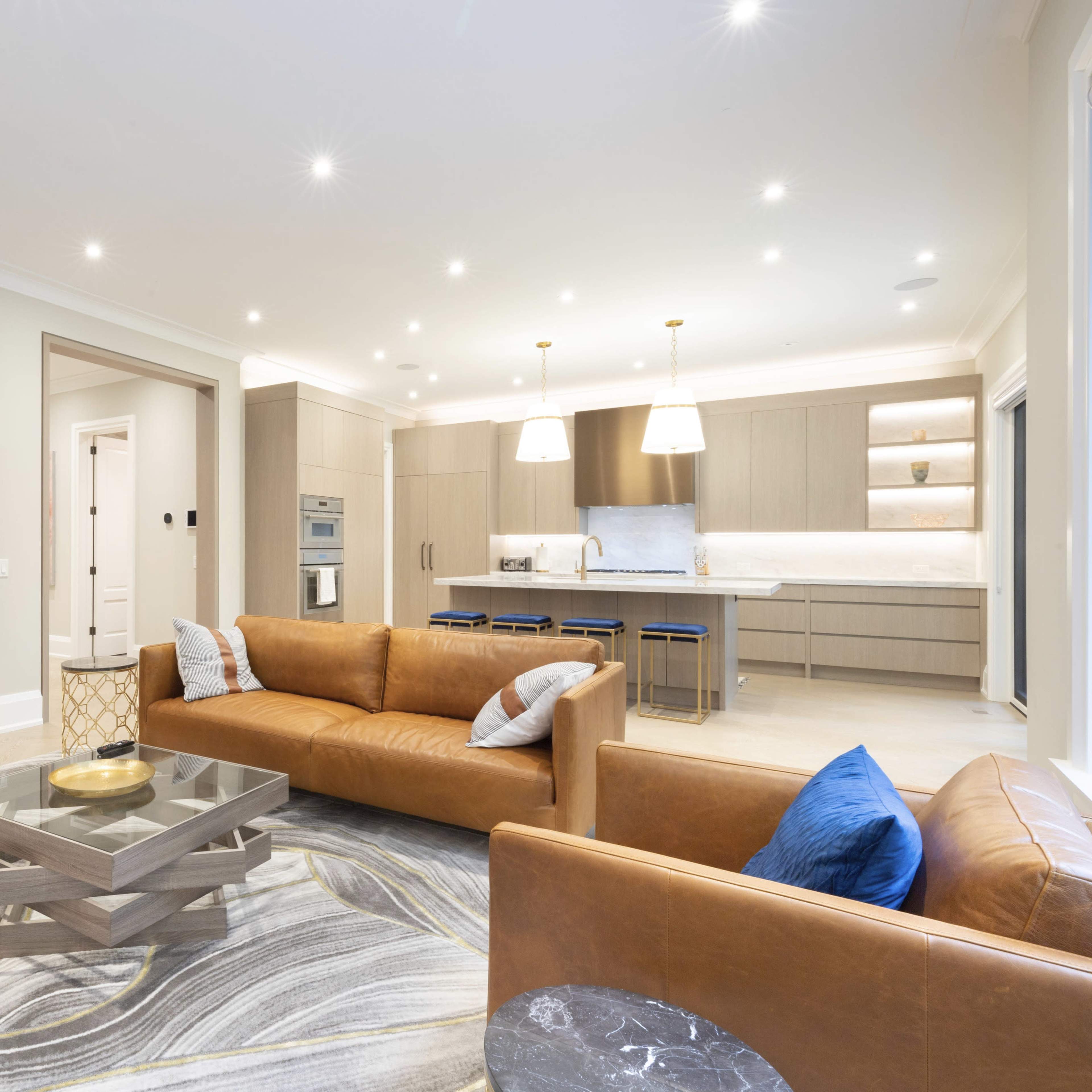 A modern living area with two leather sofas, a glass coffee table, and a large abstract painting on the wall, leading into an open kitchen with light-colored cabinetry.