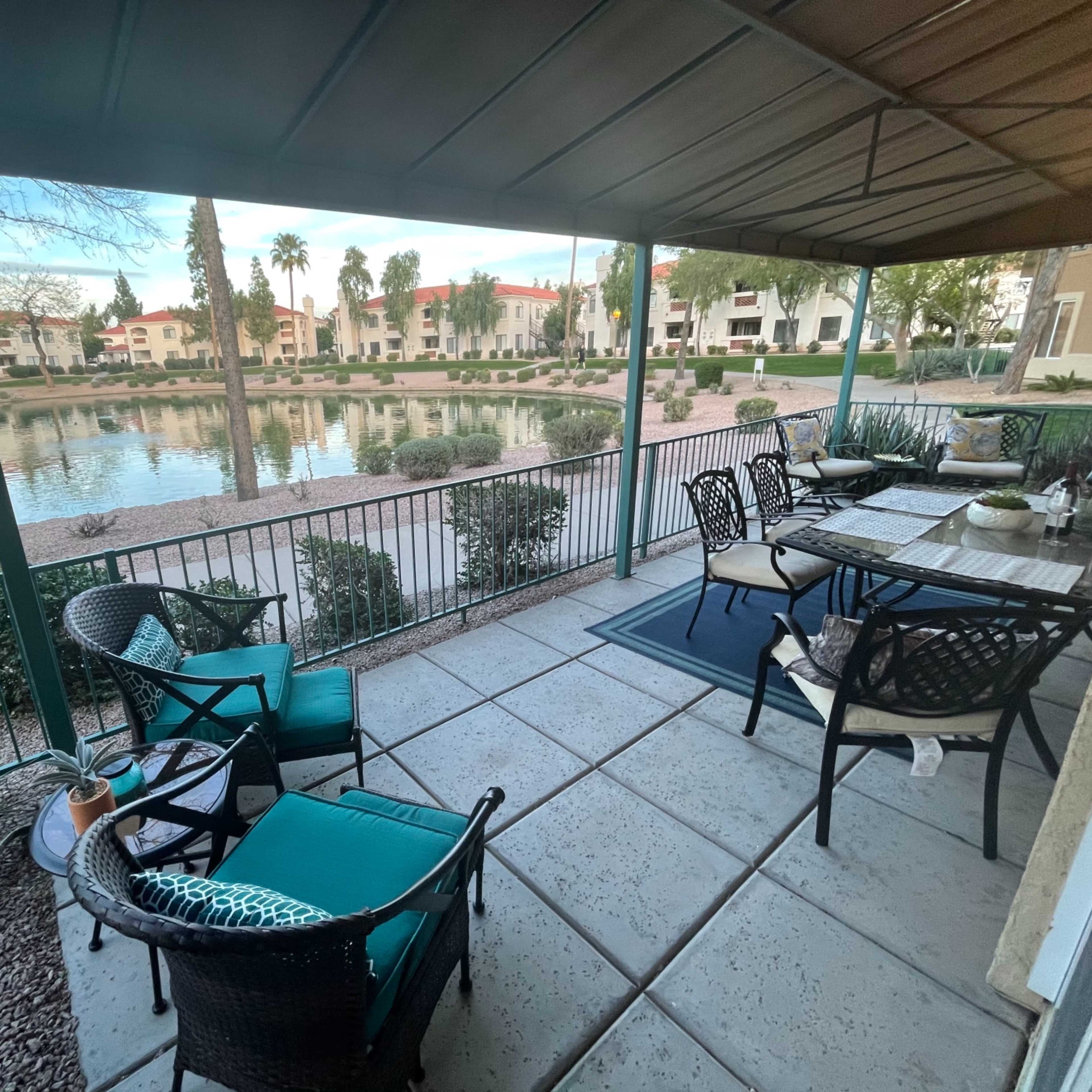 A covered patio with a table and chairs overlooks a calm pond surrounded by landscaped greenery and apartment buildings.