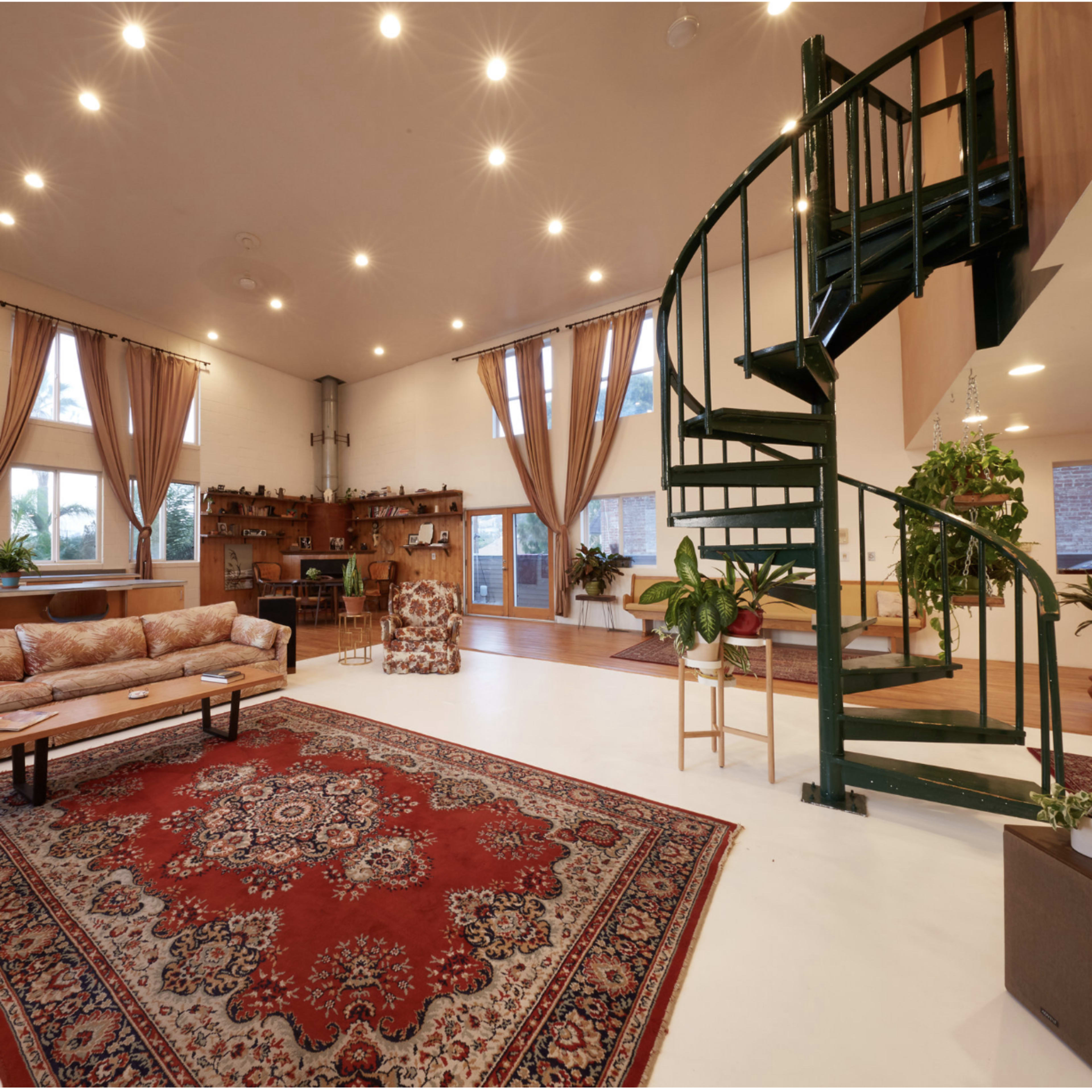 A spacious living room with large windows, a patterned rug, a spiral staircase, and several potted plants.