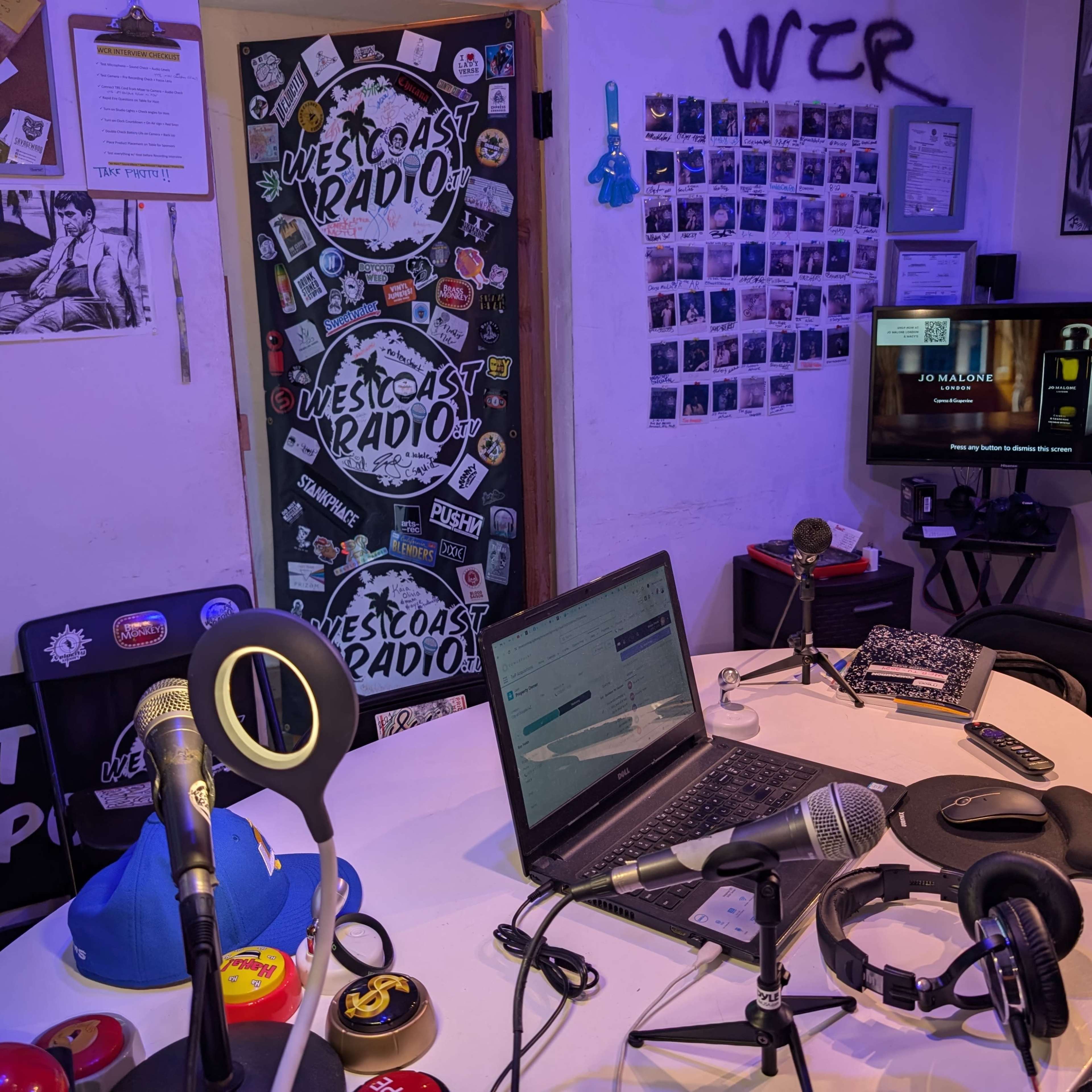A small, cluttered radio studio with microphones, a laptop, a television displaying content, and various stickers on the walls.