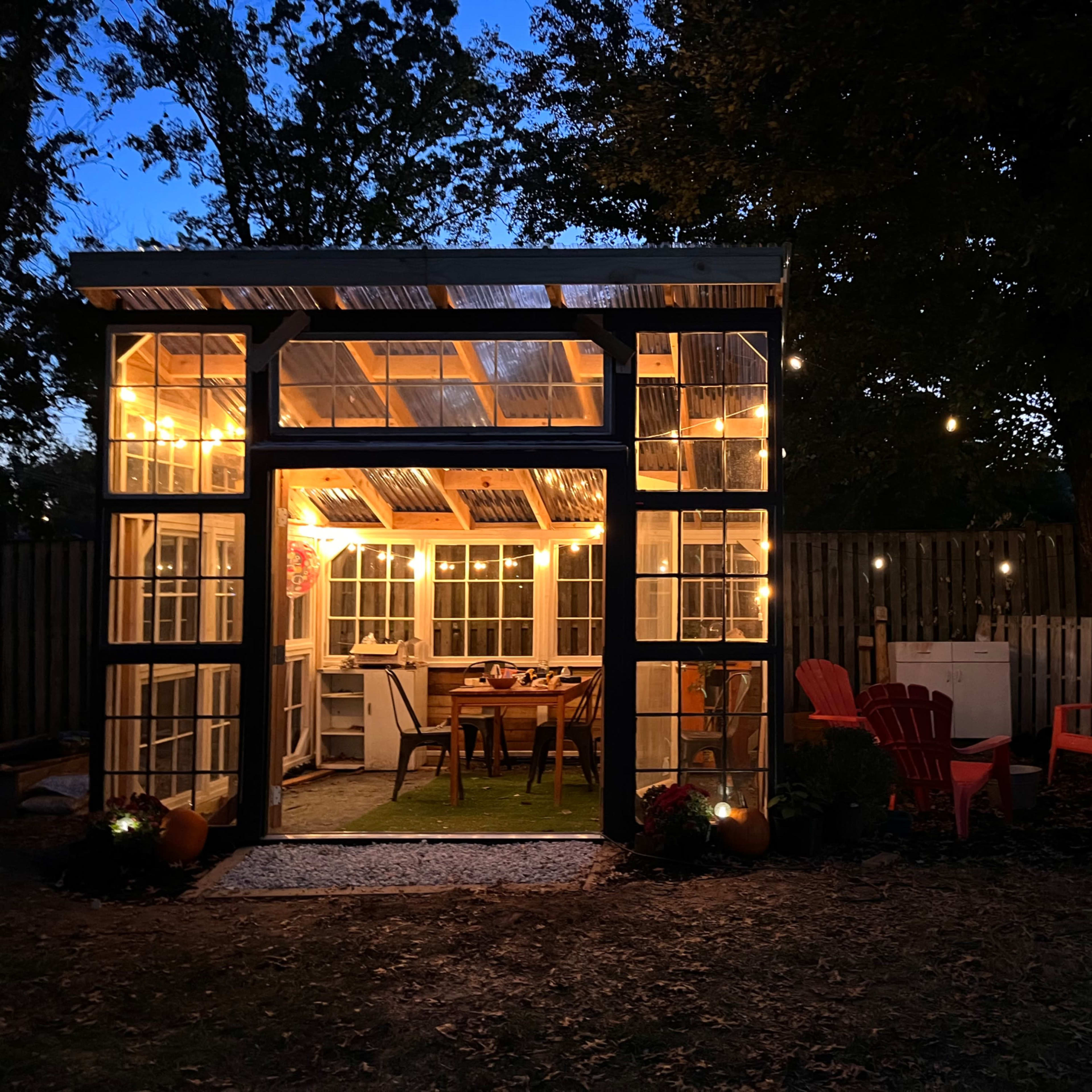 A glass greenhouse with warm lights is illuminated at dusk, surrounded by red outdoor chairs and a wooden fence.