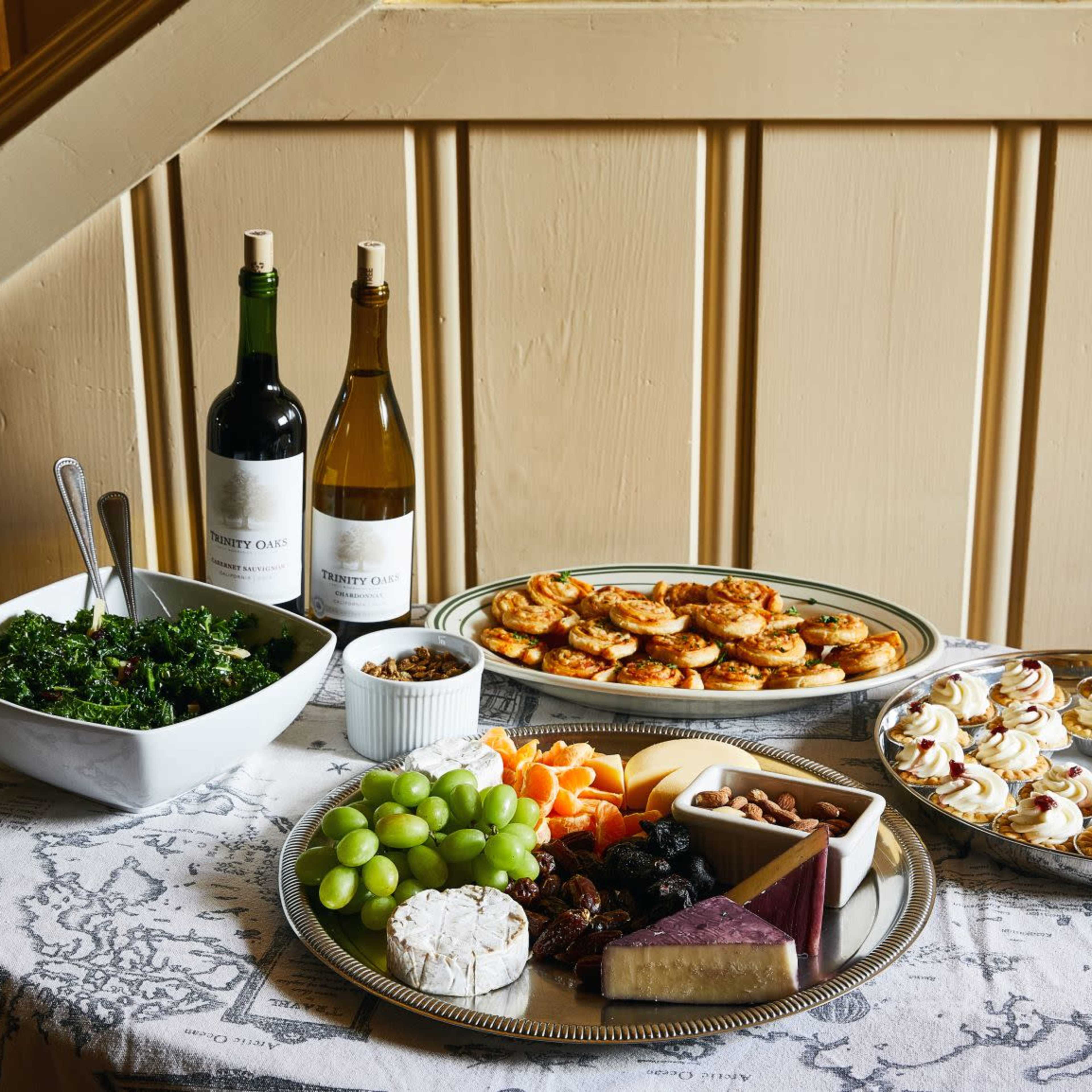 A table is set with a variety of foods, including salads, cheeses, fruits, and pastries, alongside bottles of red and white wine.