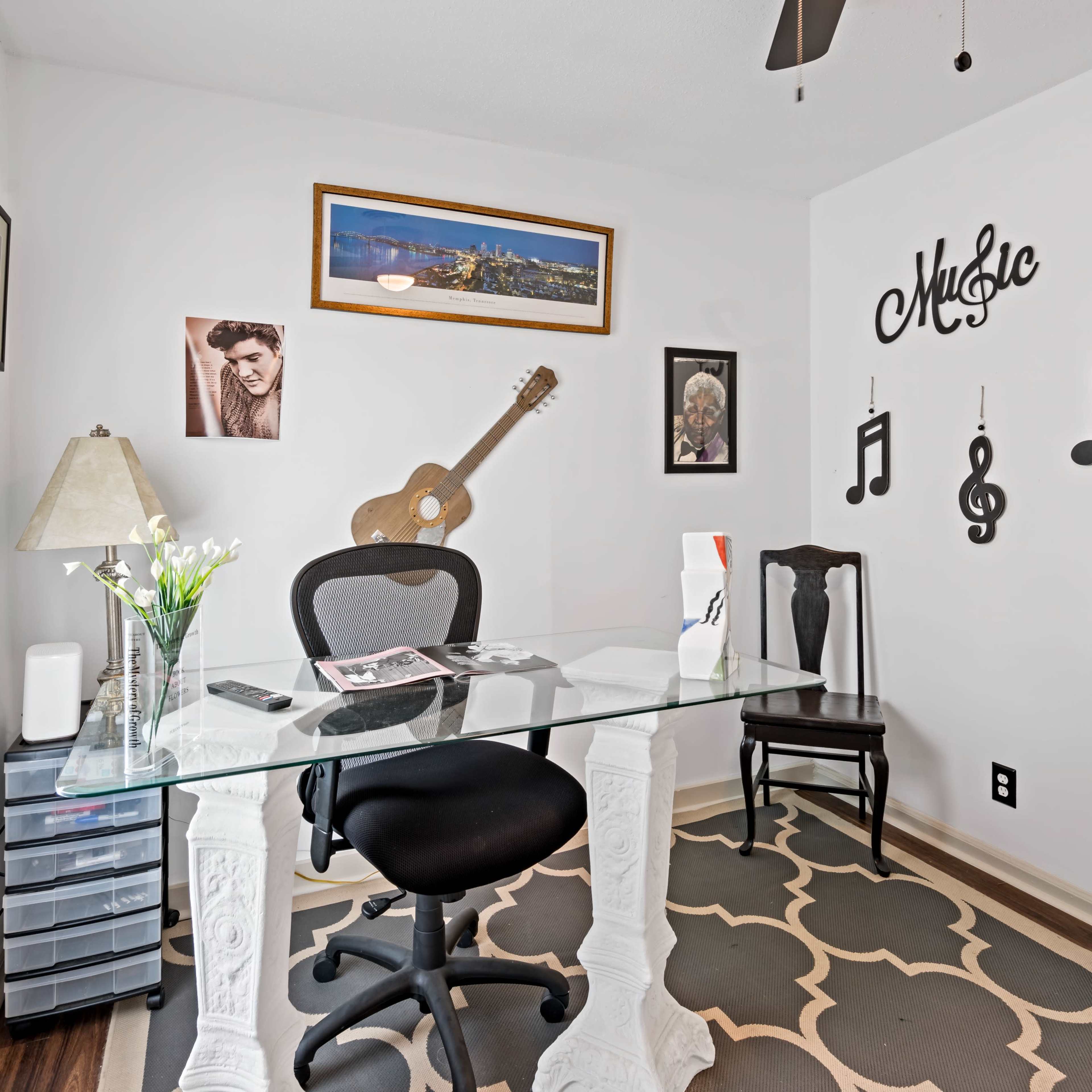 A home office features a glass desk, two chairs, a lamp, and musical decor against a white wall.