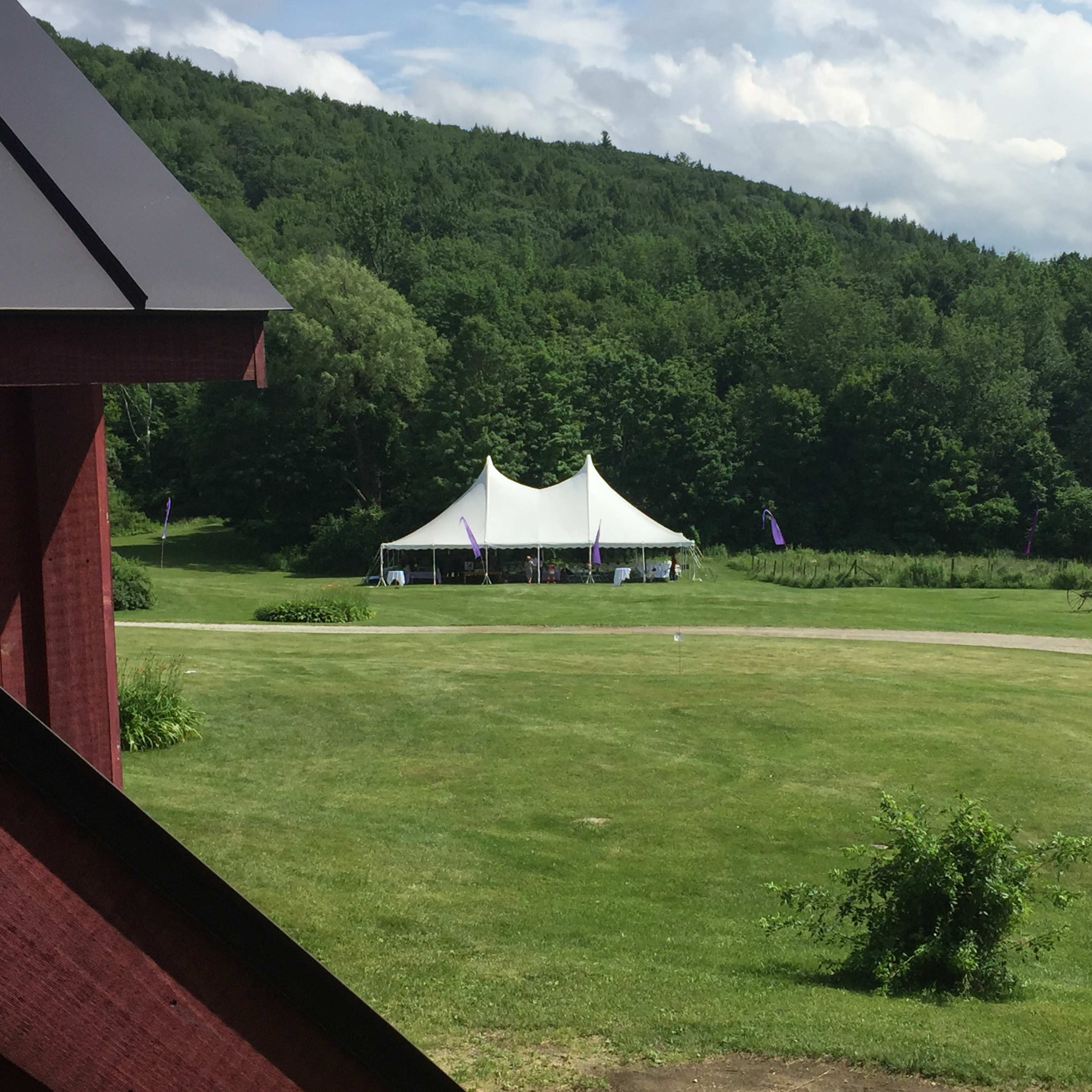 A large white tent is set up on a grassy area surrounded by trees and hills, with a few people walking nearby.