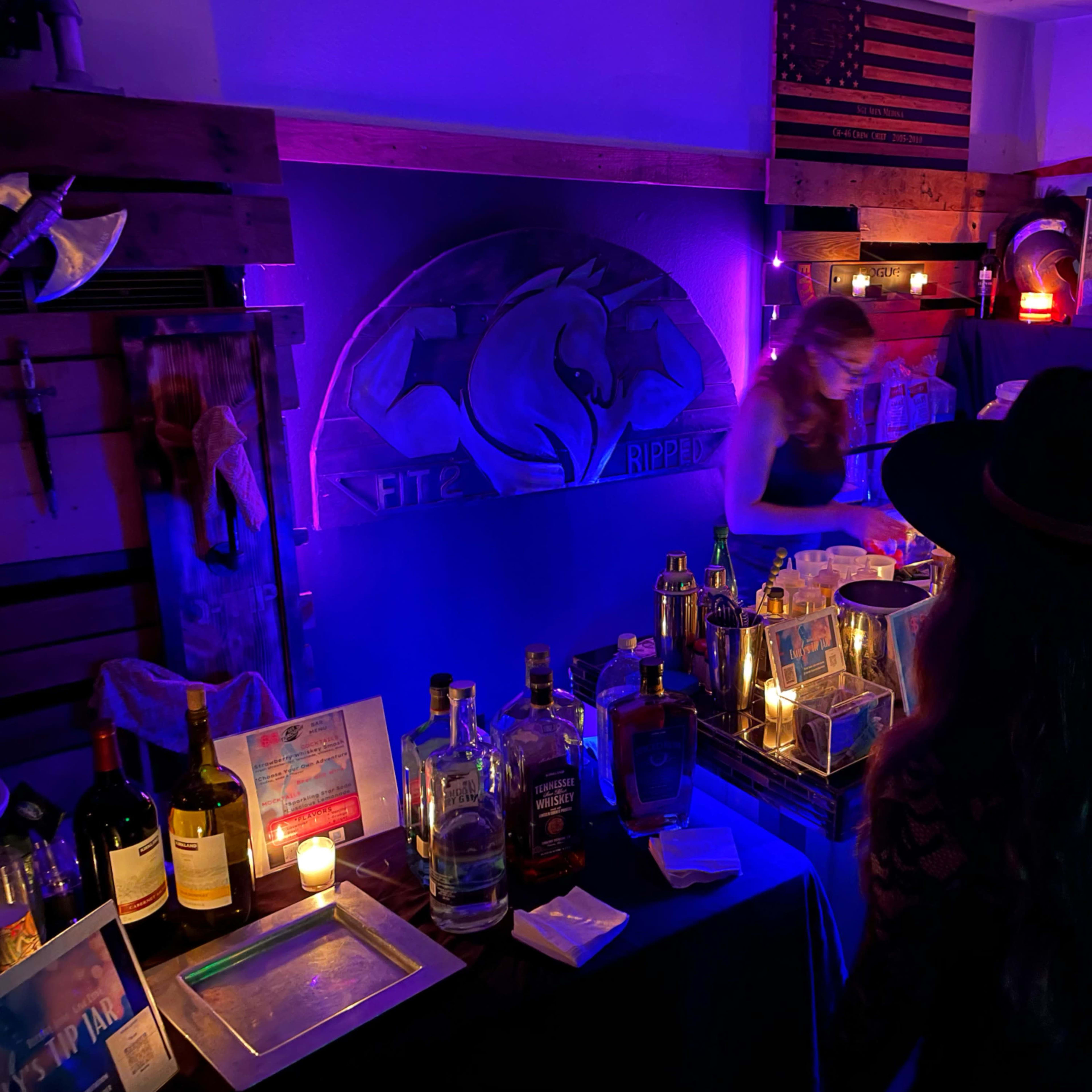 A dimly lit bar area decorated with purple lighting, featuring various bottles of alcohol and two women standing near the counter.