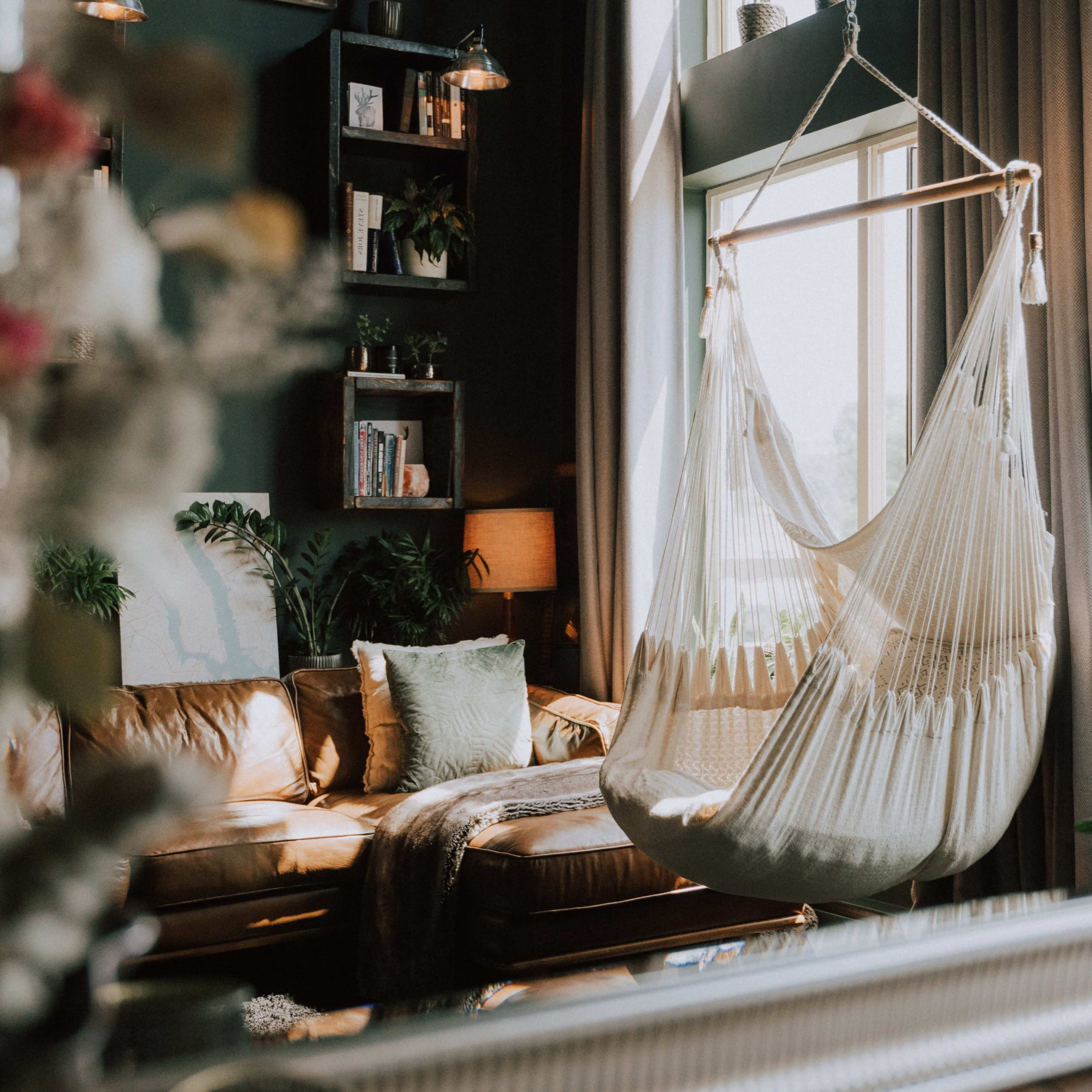 A cozy living room featuring a hammock chair hanging beside a leather sofa, with large windows allowing natural light to fill the space.