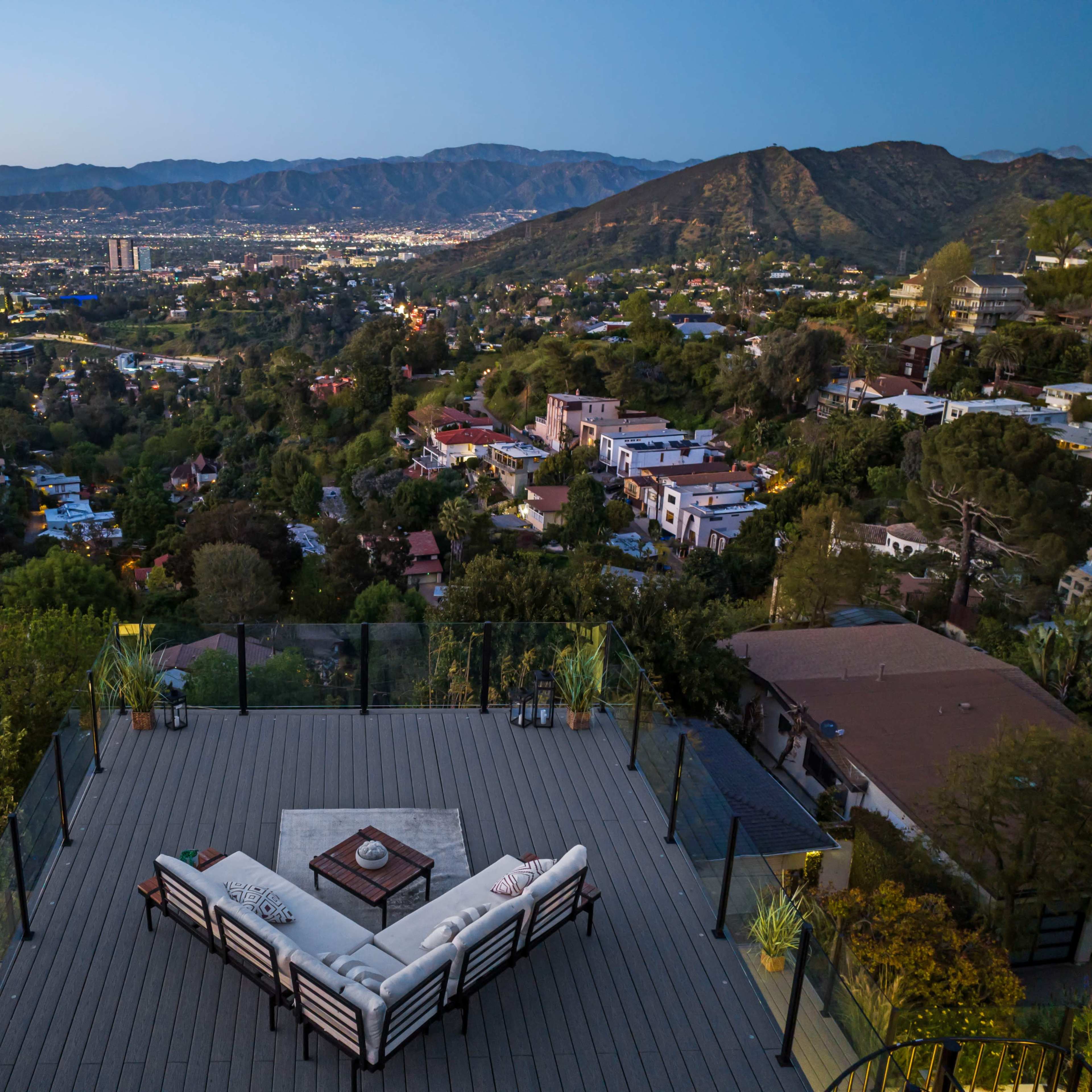 A spacious outdoor terrace with a sectional sofa overlooks a valley filled with homes and mountains at dusk.