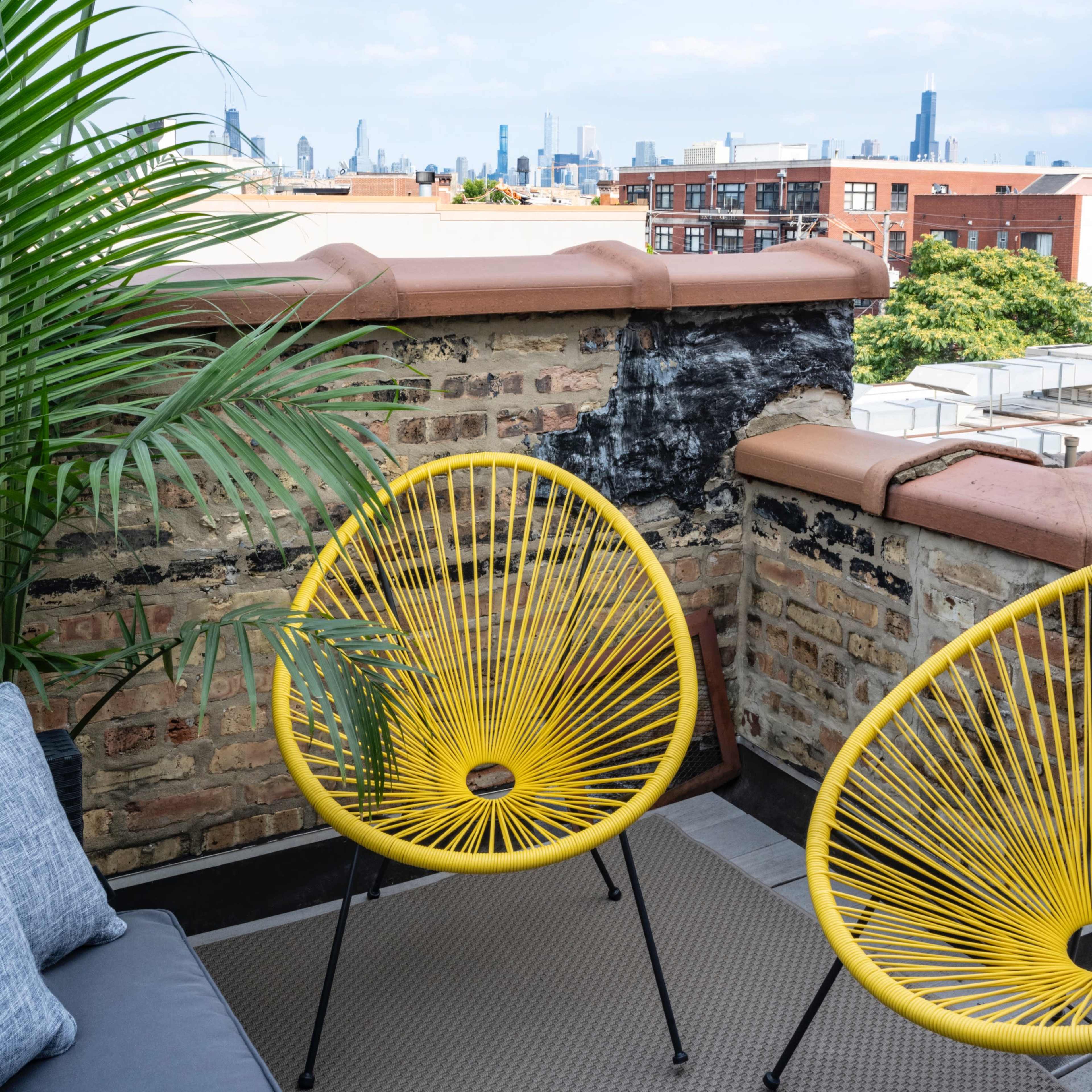 Two yellow chairs on a rooftop patio with a city skyline in the background and a potted plant nearby.
