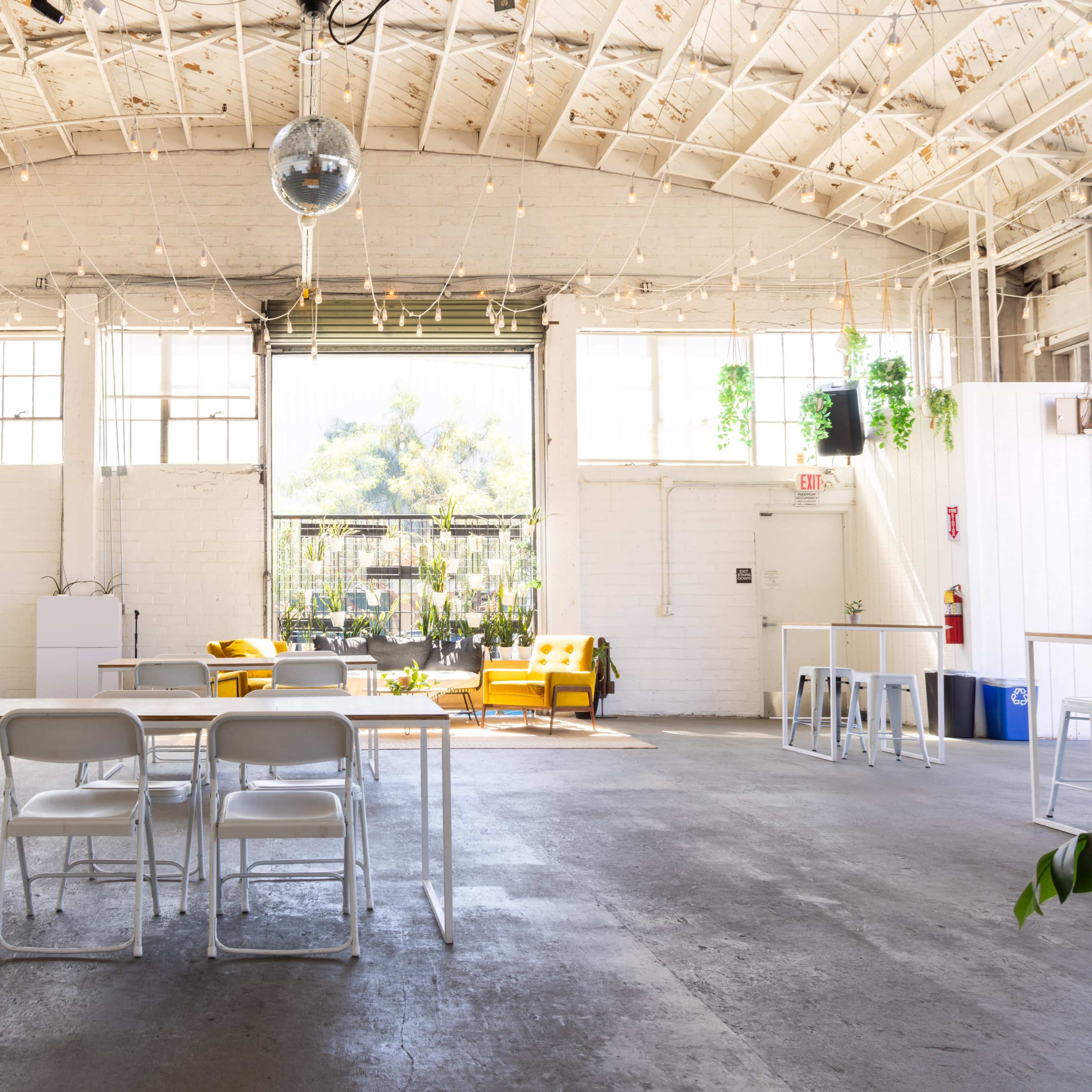 An open, bright room with large windows, featuring simple furniture, plants, and a disco ball hanging from the ceiling.