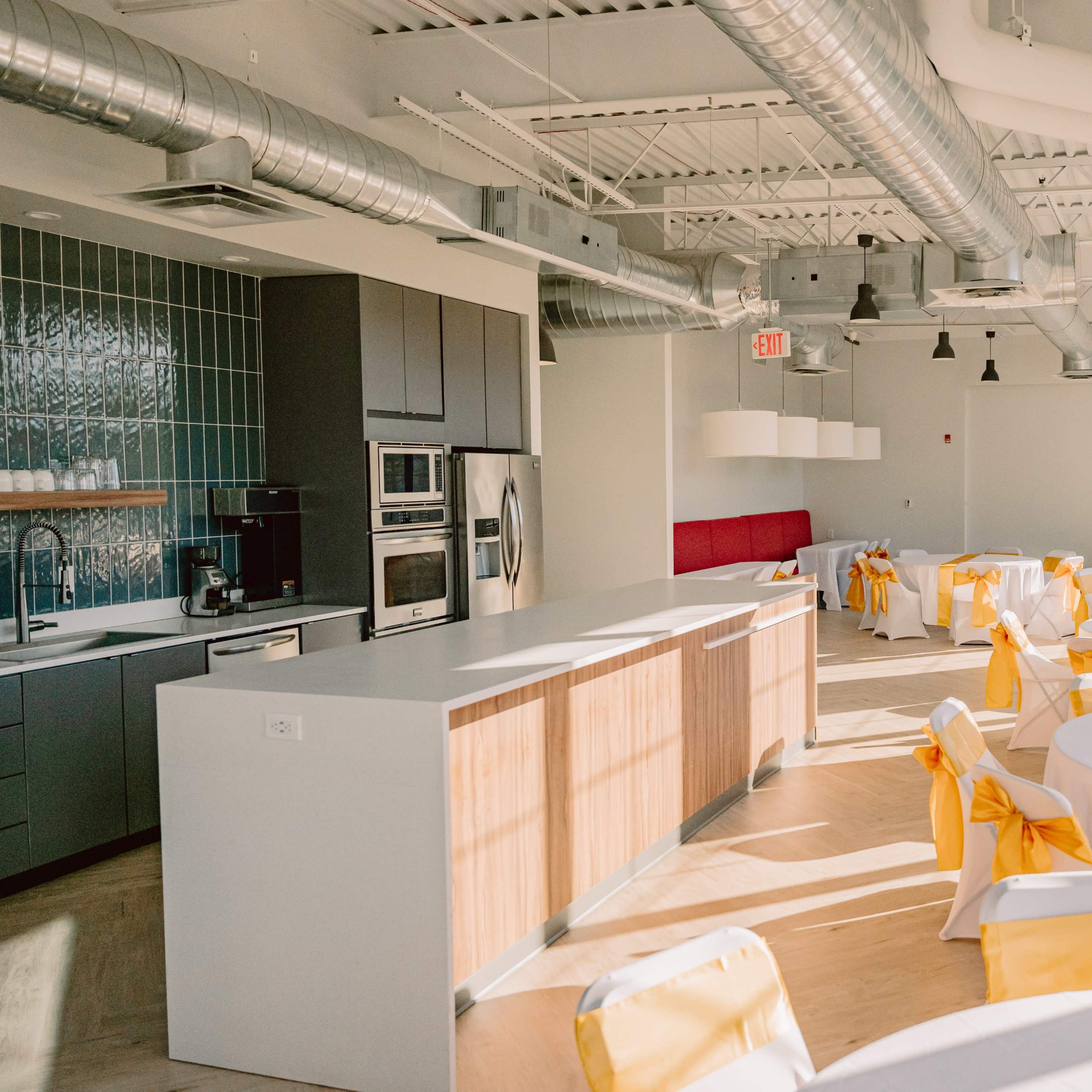 A bright event space featuring round tables with yellow table runners and chair sashes, along with modern kitchen appliances in the background.