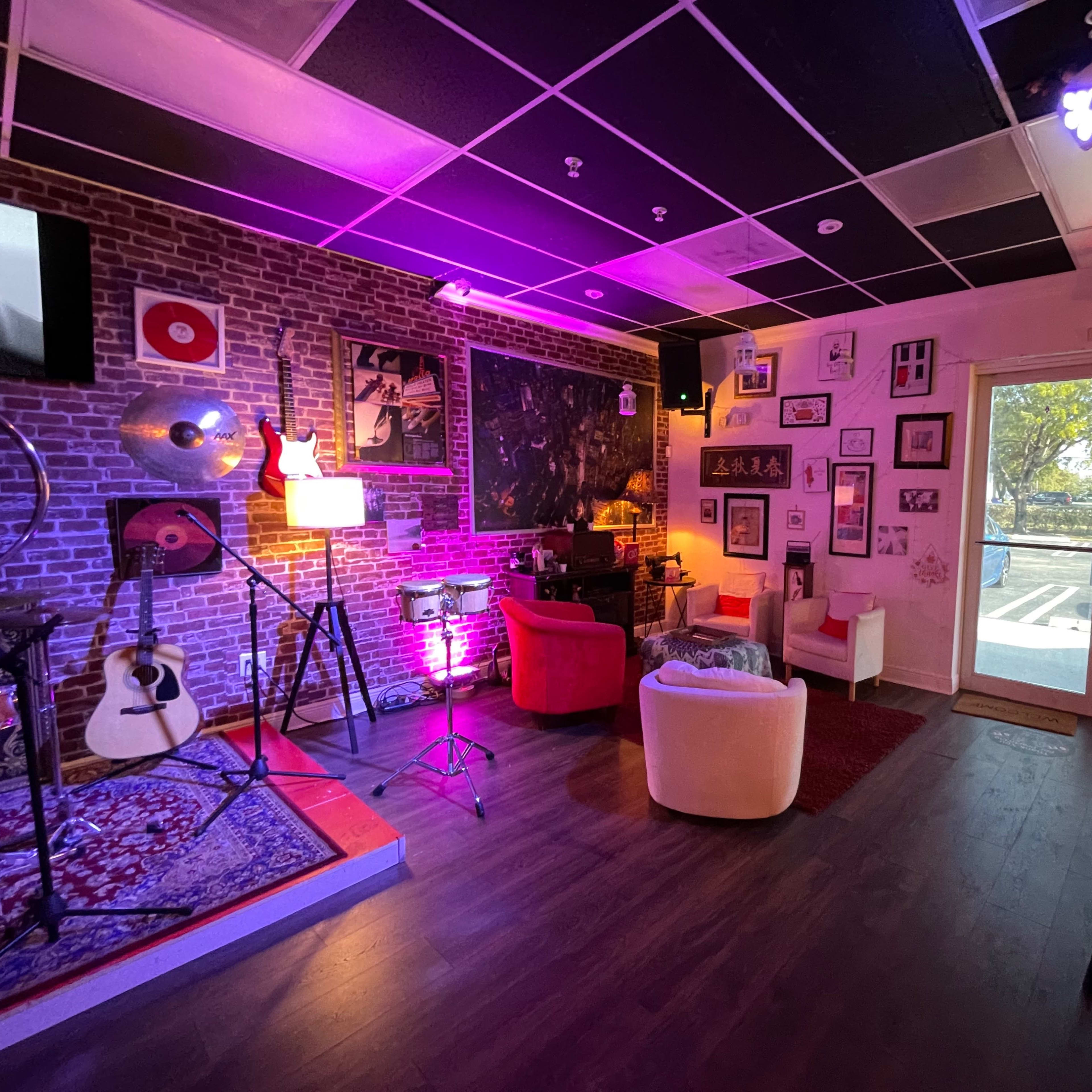A small, cozy lounge area with a brick wall, musical instruments, and various wall decorations, illuminated by purple and white lights.