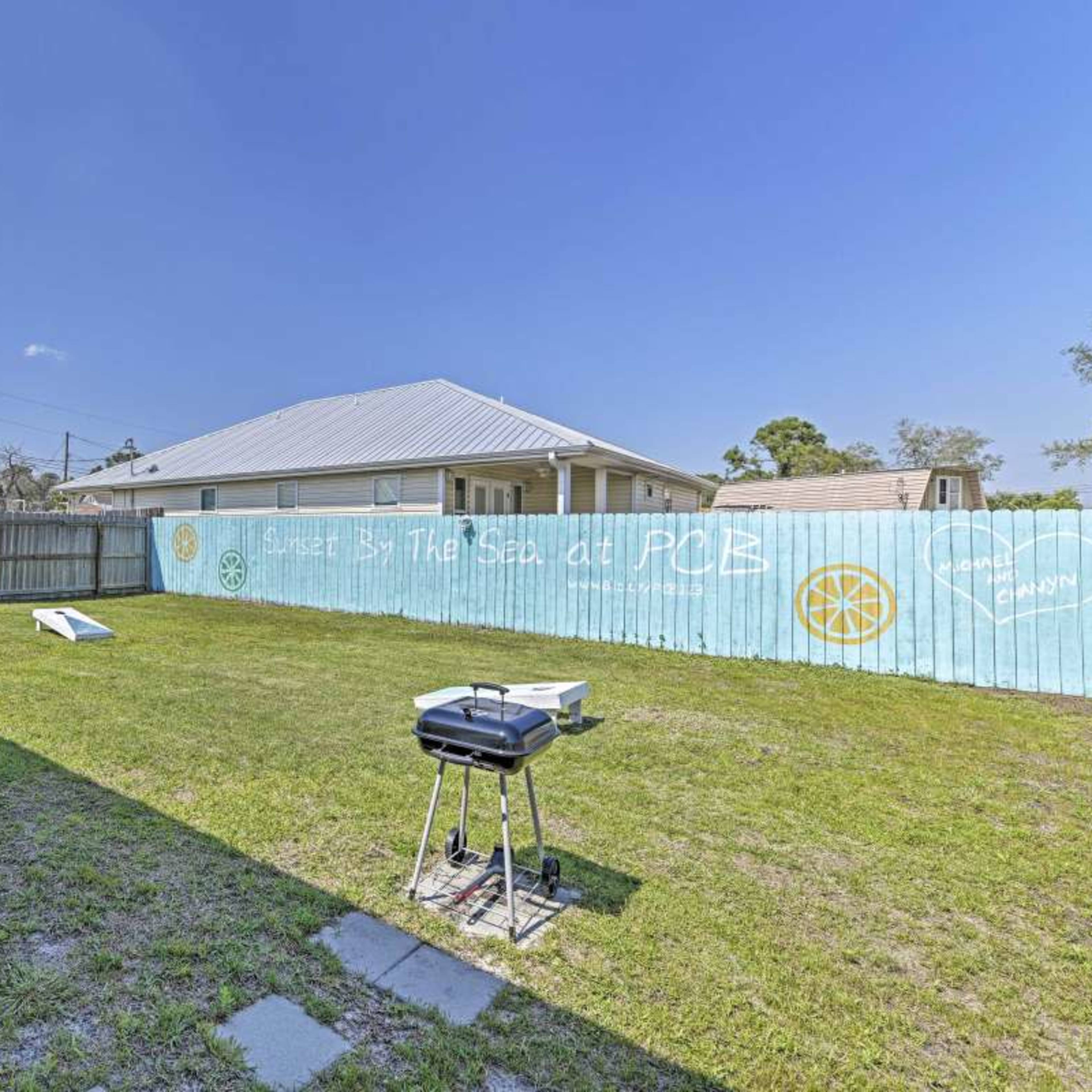 A backyard features a green lawn with a grill and a cornhole game set up next to a wooden fence with a mural.