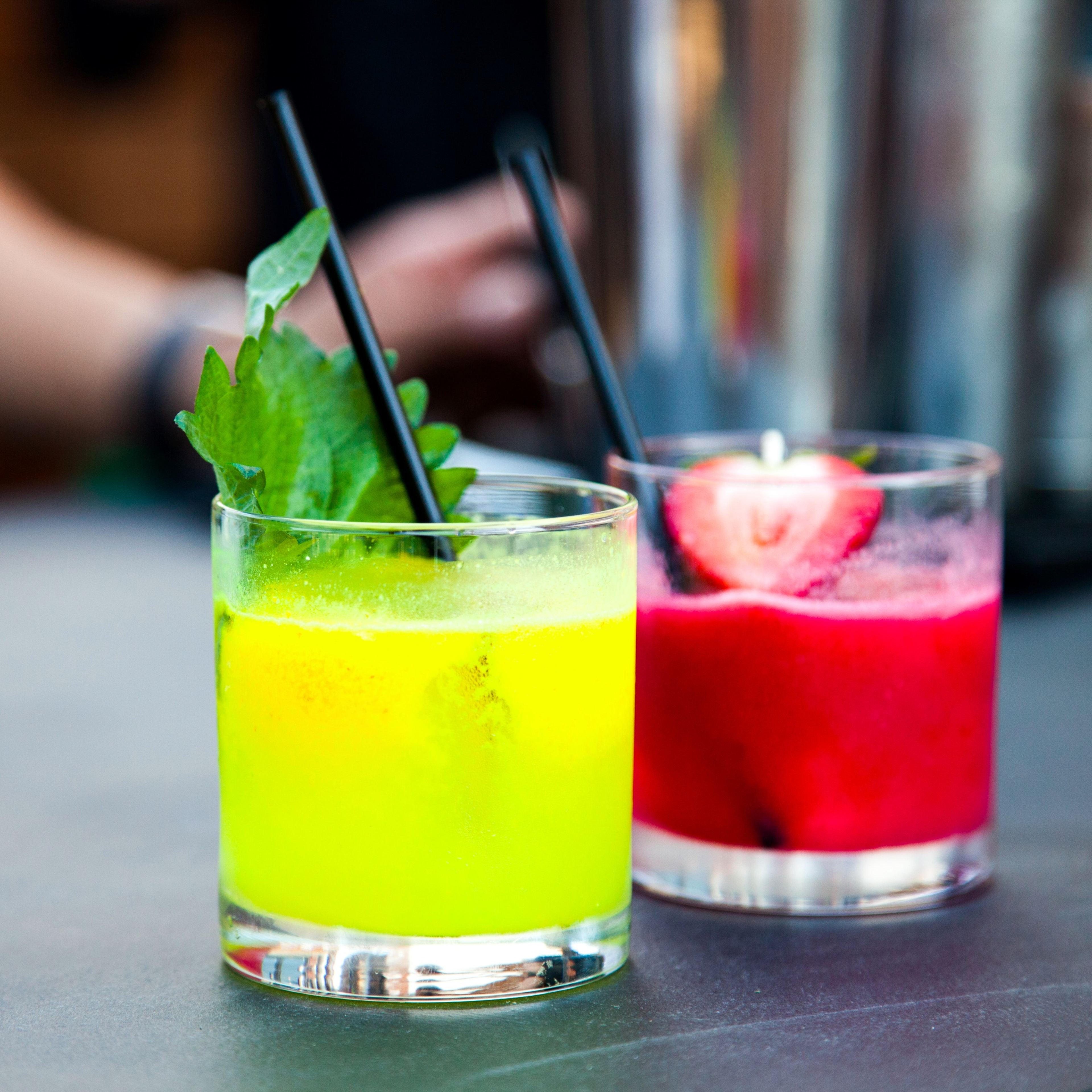 Two colorful cocktails, one bright green with mint leaves and the other red with a strawberry, sit on a table with black straws.