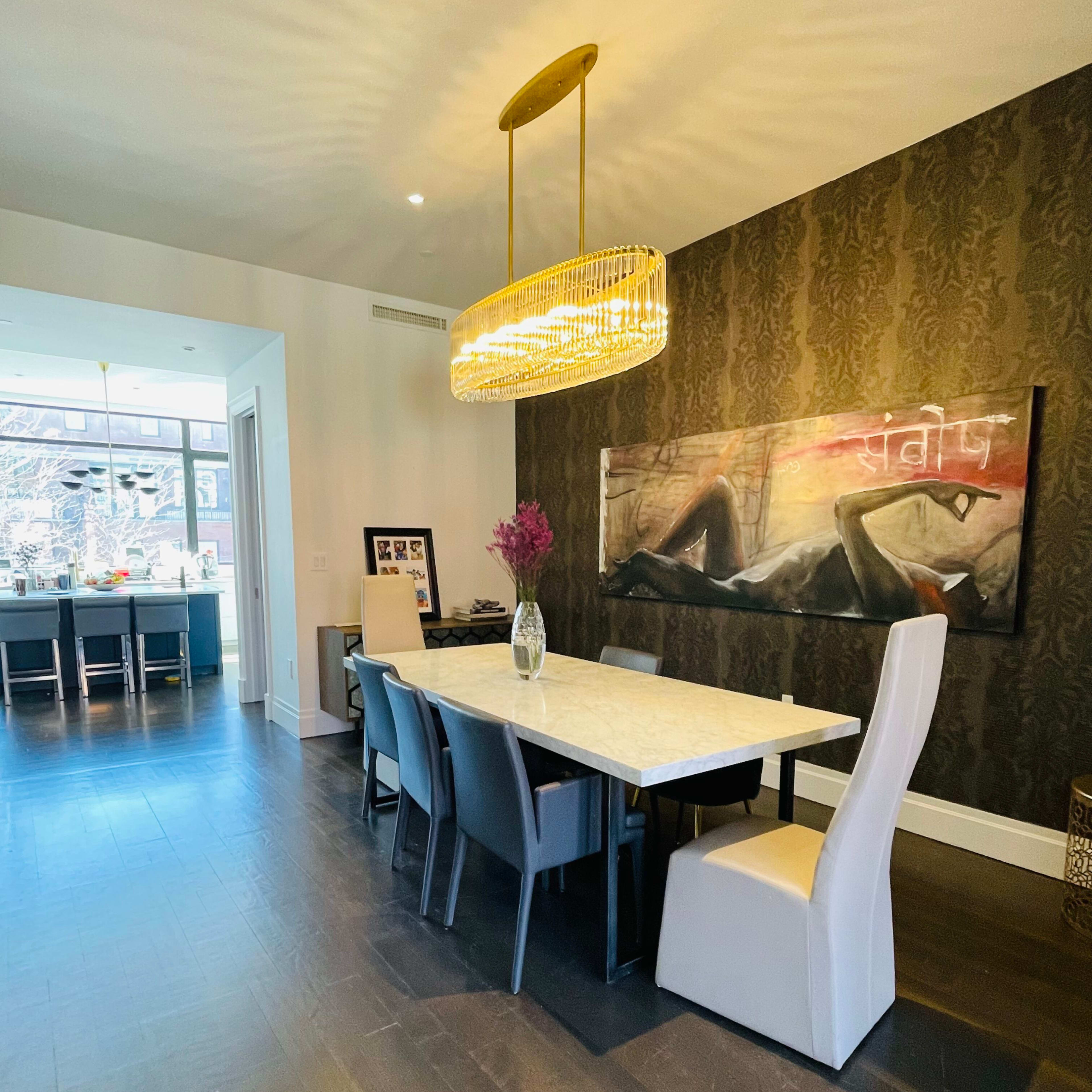 A modern dining area features a long white table surrounded by black and white chairs, with a large artwork on the wall and a pendant light overhead.