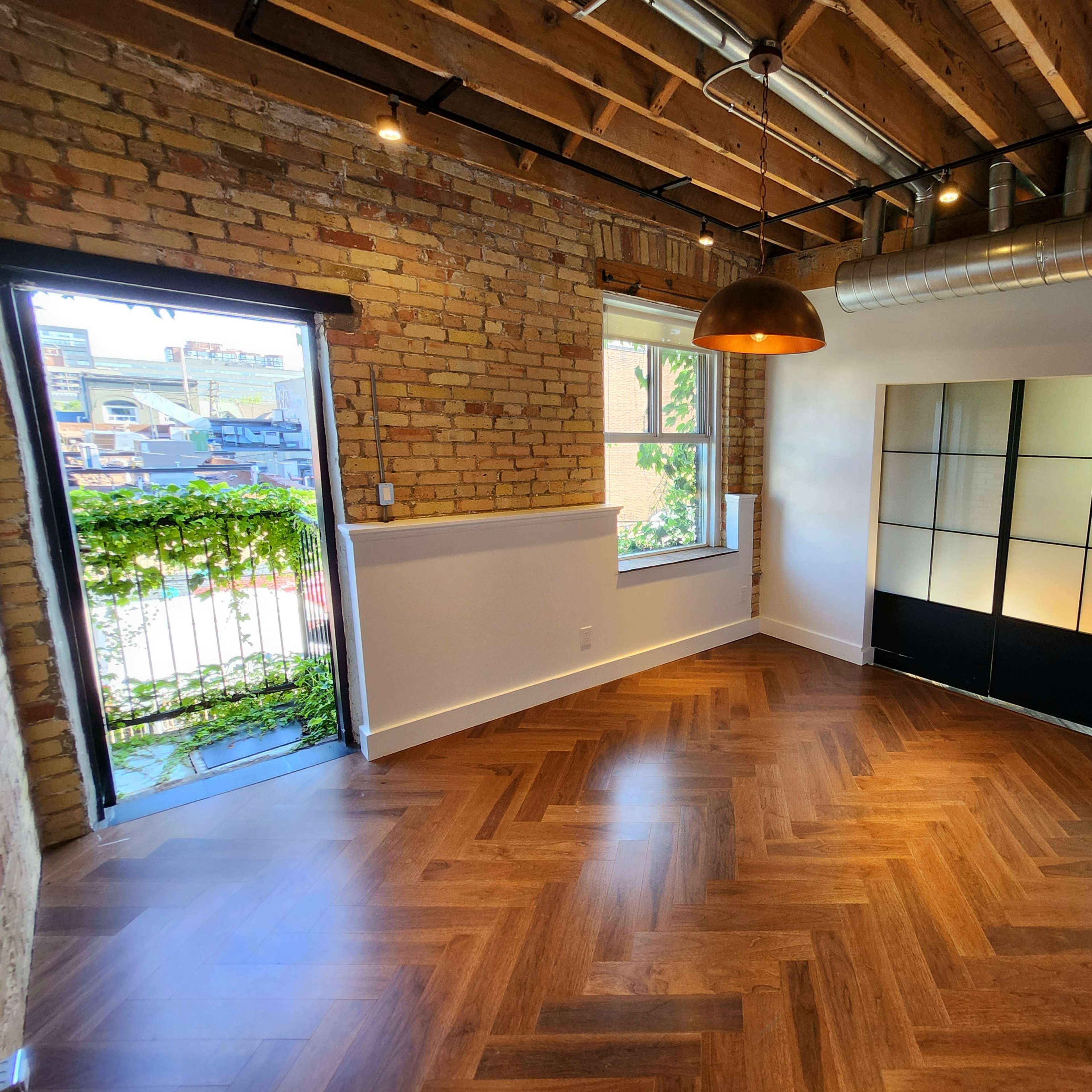 A room with exposed brick walls, wooden flooring in a herringbone pattern, a large window facing a balcony, and a decorative black partition.