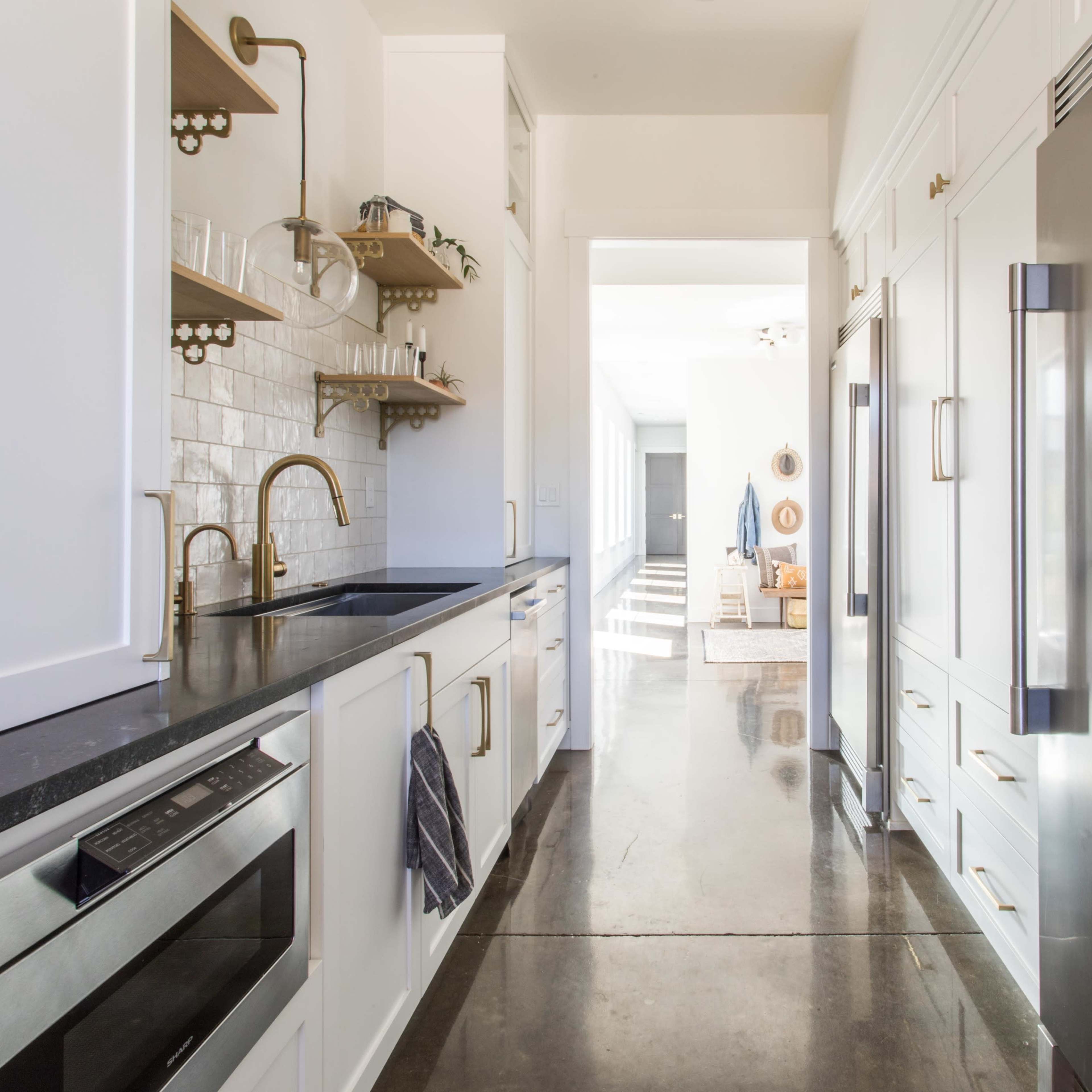 A narrow kitchen features modern cabinetry, a stainless steel refrigerator, and an adjoining open hallway leading to a well-lit living area.