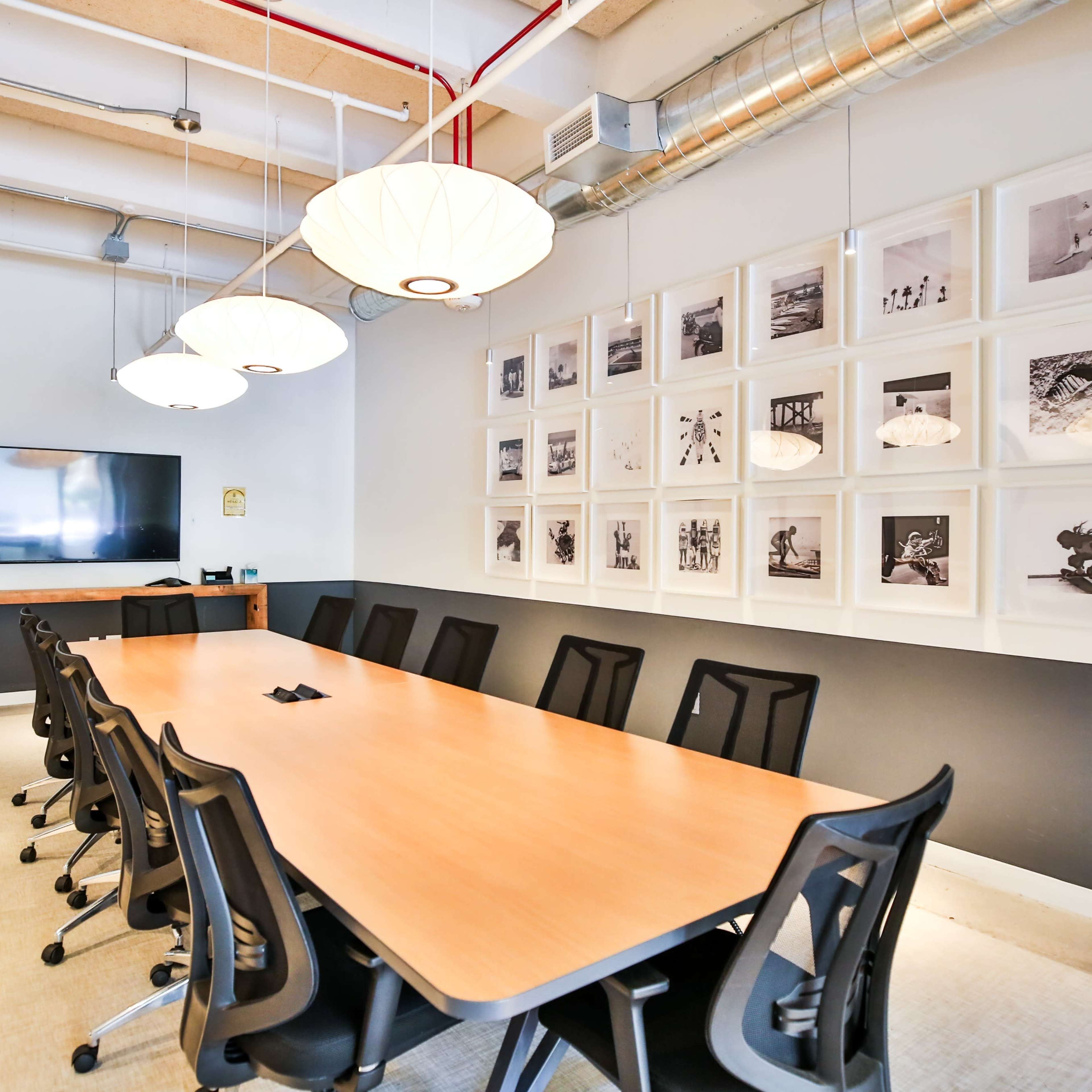 A conference room features a long wooden table surrounded by black chairs, with framed photographs on the wall and a television mounted opposite the table.