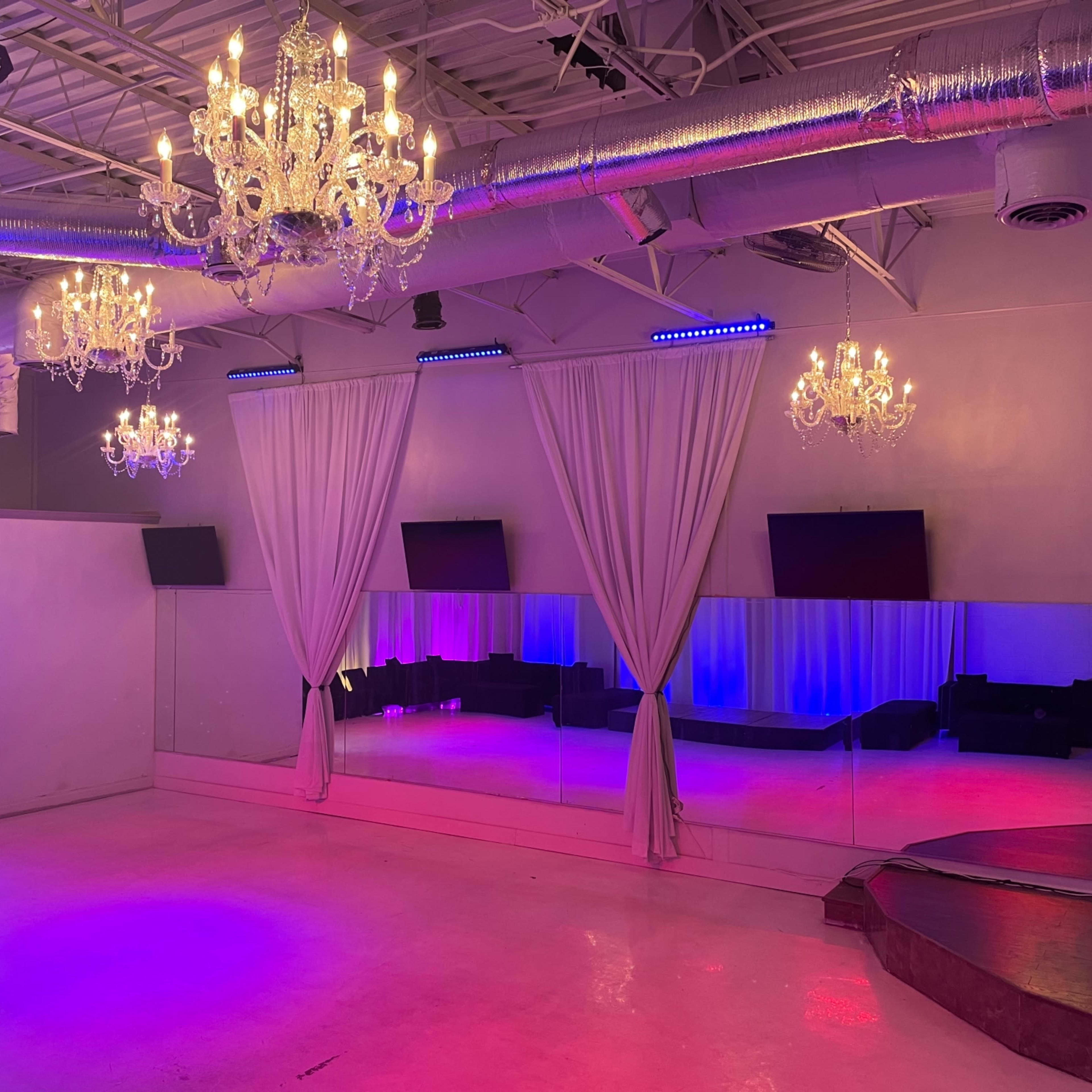 A spacious event venue with mirrored walls, chandeliers, and subtle colored lighting.