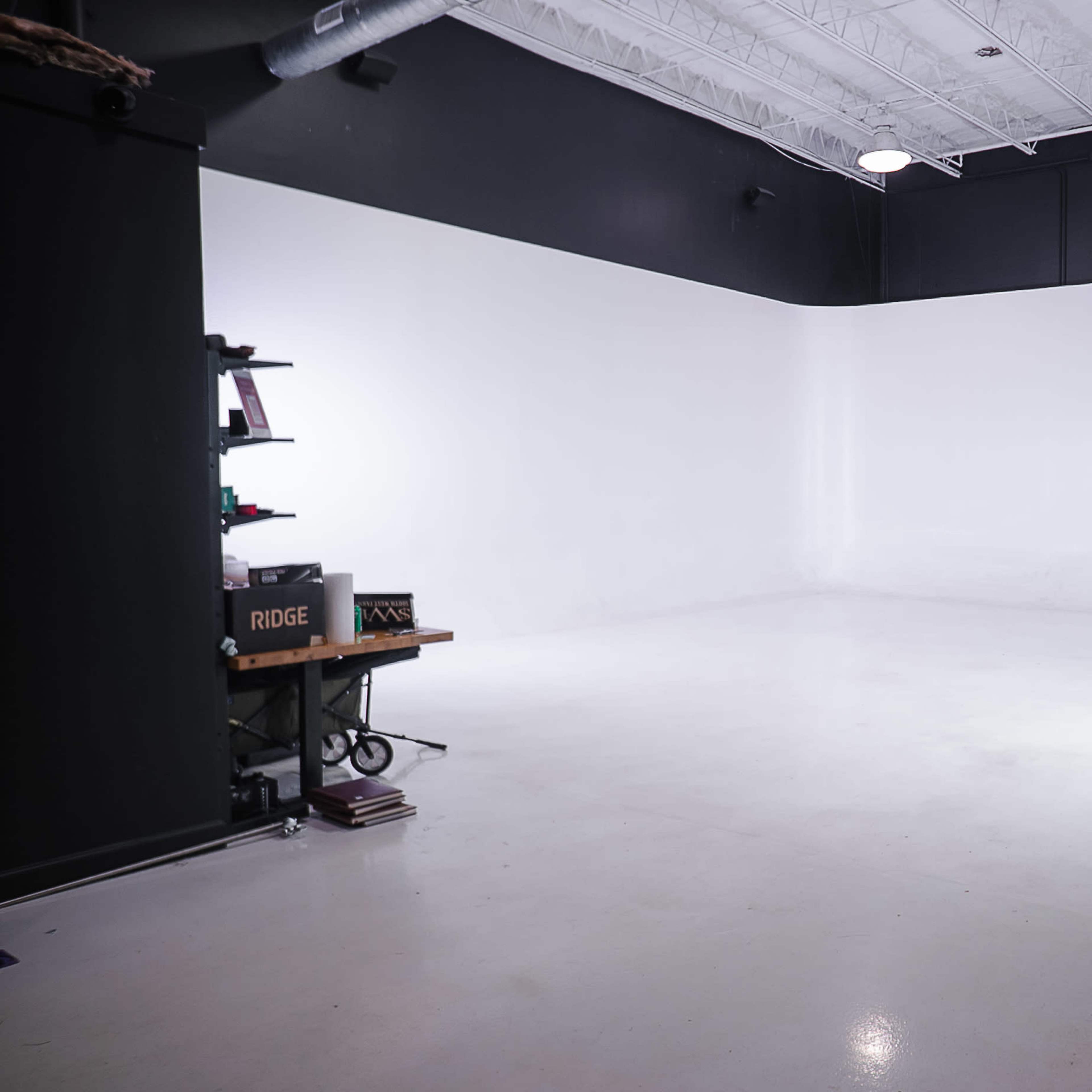 An empty photography studio with a white backdrop and minimal equipment along one wall.